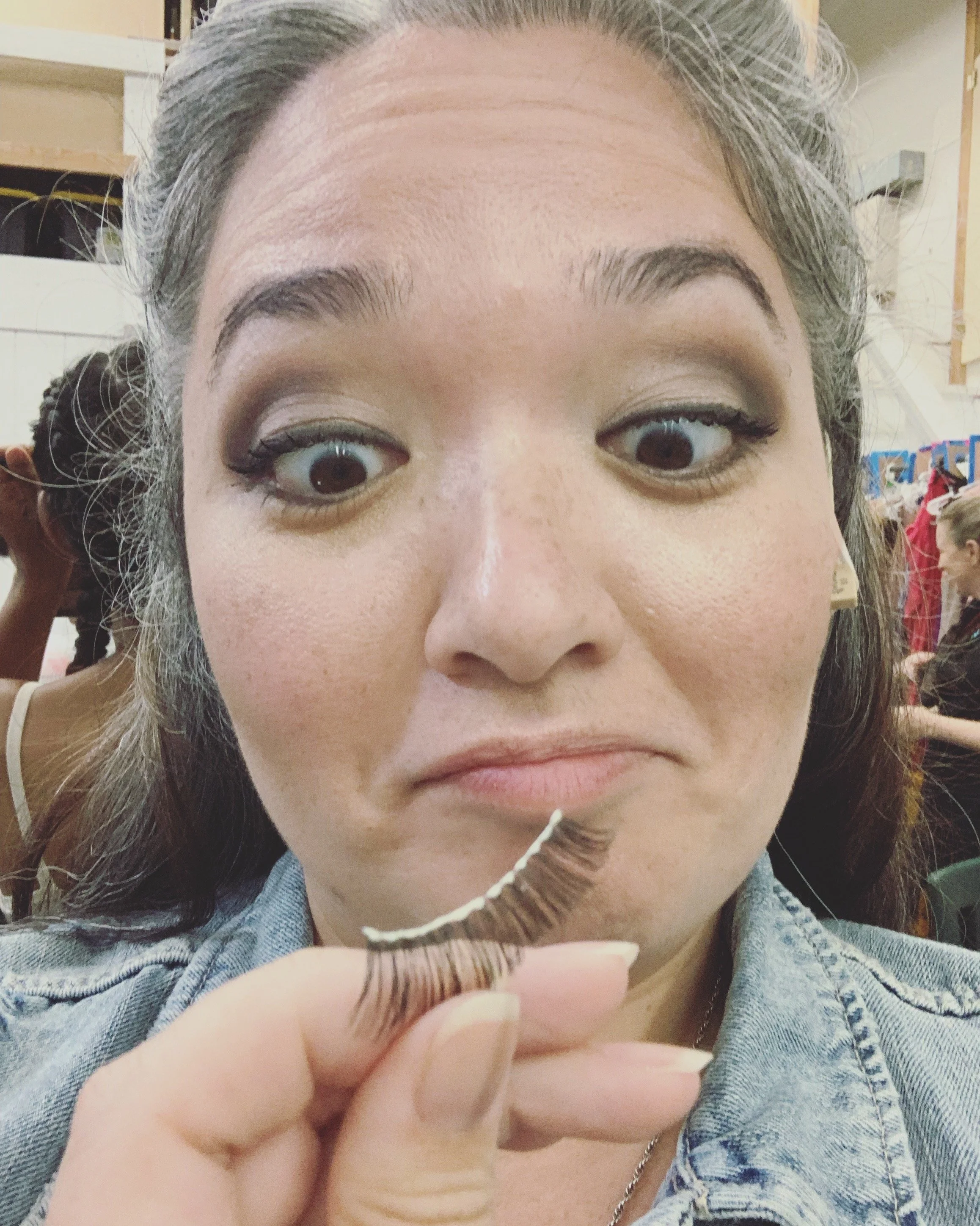 CHILDREN OF EDEN a Broadway musical by I'm A Bright Kid Foundation at Paliku Theatre. A woman with makeup holding a false eyelash in front of her lips, in a room with people in the background.