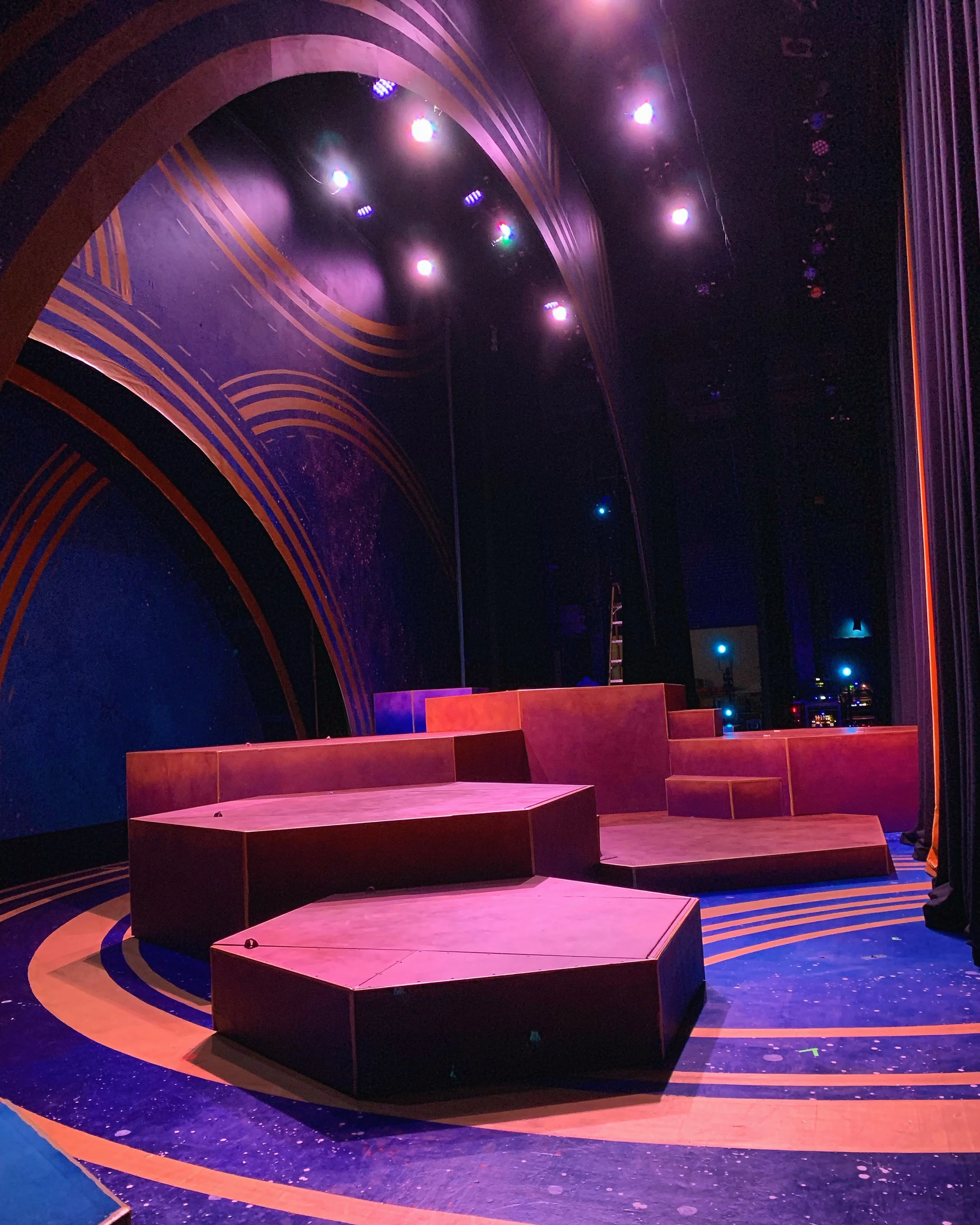 CHILDREN OF EDEN a Broadway musical by I'm A Bright Kid Foundation. The stage of a theater under purple and pink stage lights, featuring geometric, multi-tiered platforms with black edges, and a backdrop with gold lines and a dark blue background.