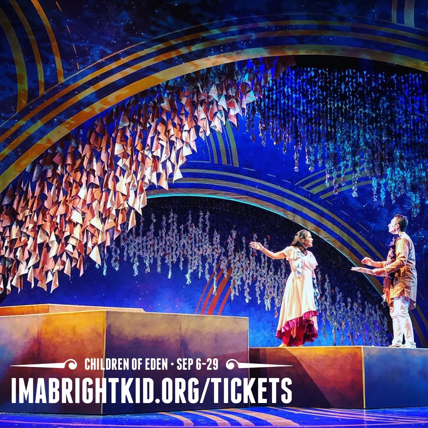 Stage scene from the play 'Children of Eden' featuring two actors, one woman in a pink dress with flowers, another young man in a brown jacket, standing under a colorful, cosmic-themed set with hanging paper decorations and golden curved arches.