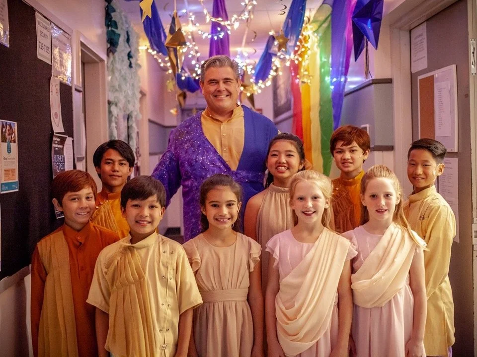 A group of children dressed in colorful dresses and traditional Indian attire with a man in a vibrant costume, celebrating a cultural or festive event indoors decorated with rainbow-colored flowers, streamers, and hanging ornaments.