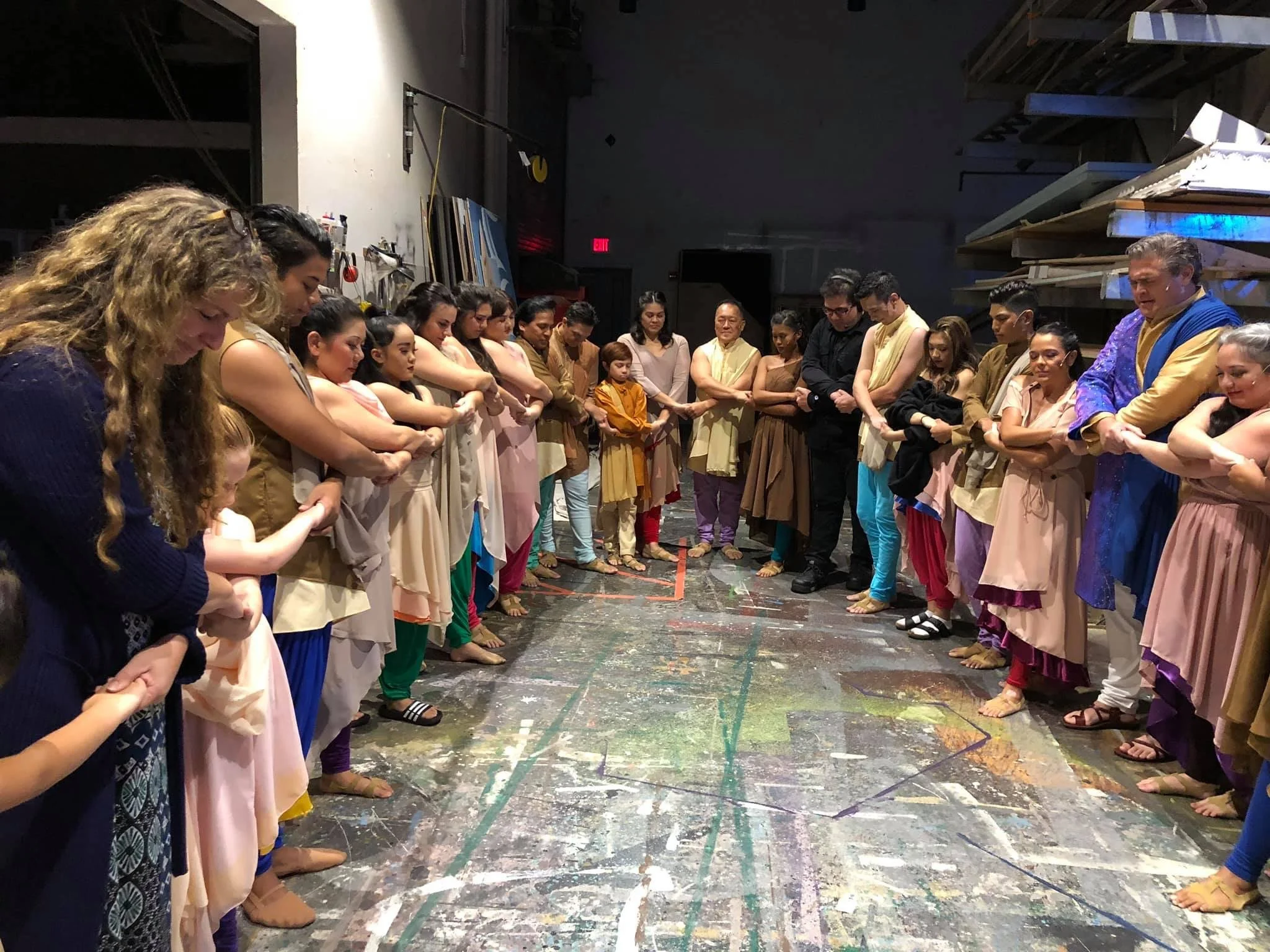 CHILDREN OF EDEN a Broadway musical by I'm A Bright Kid Foundation at Paliku Theatre. Group of people standing in a circle with arms crossed, inside an indoor studio or warehouse, some dressed in traditional Indian attire.