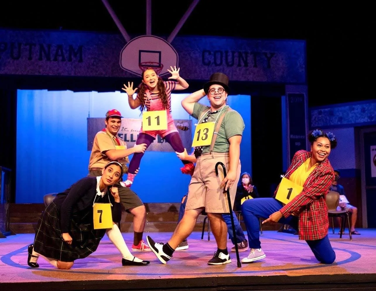 THE 25TH ANNUAL PUTNAM COUNTY SPELLING BEE a comedy musical. The cast of a play performing on stage, dressed in colorful costumes with numbered tags, in front of a backdrop with a basketball hoop and signs indicating a community or school setting.