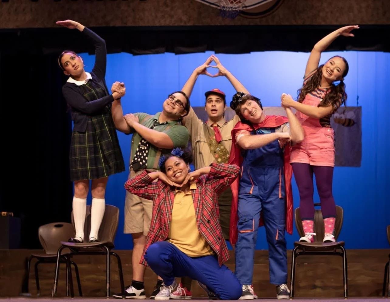 THE 25TH ANNUAL PUTNAM COUNTY SPELLING BEE a comedy musical at Manoa Valley Theatre in Honolulu, Hawaii. A group of five performers on stage, posing playfully with two placed on chairs, all in colorful costumes with a blue background.