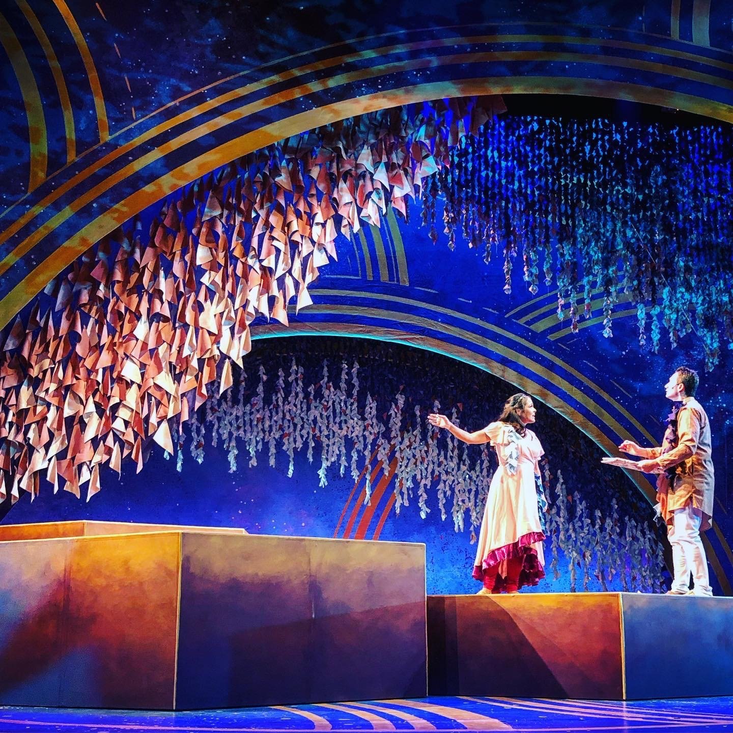 CHILDREN OF EDEN a Broadway musical by I'm A Bright Kid Foundation at Paliku. Two actors performing on a colorful stage with elaborate arch designs and hanging paper decorations, one woman in a dress extending her arm towards a man holding a tray.