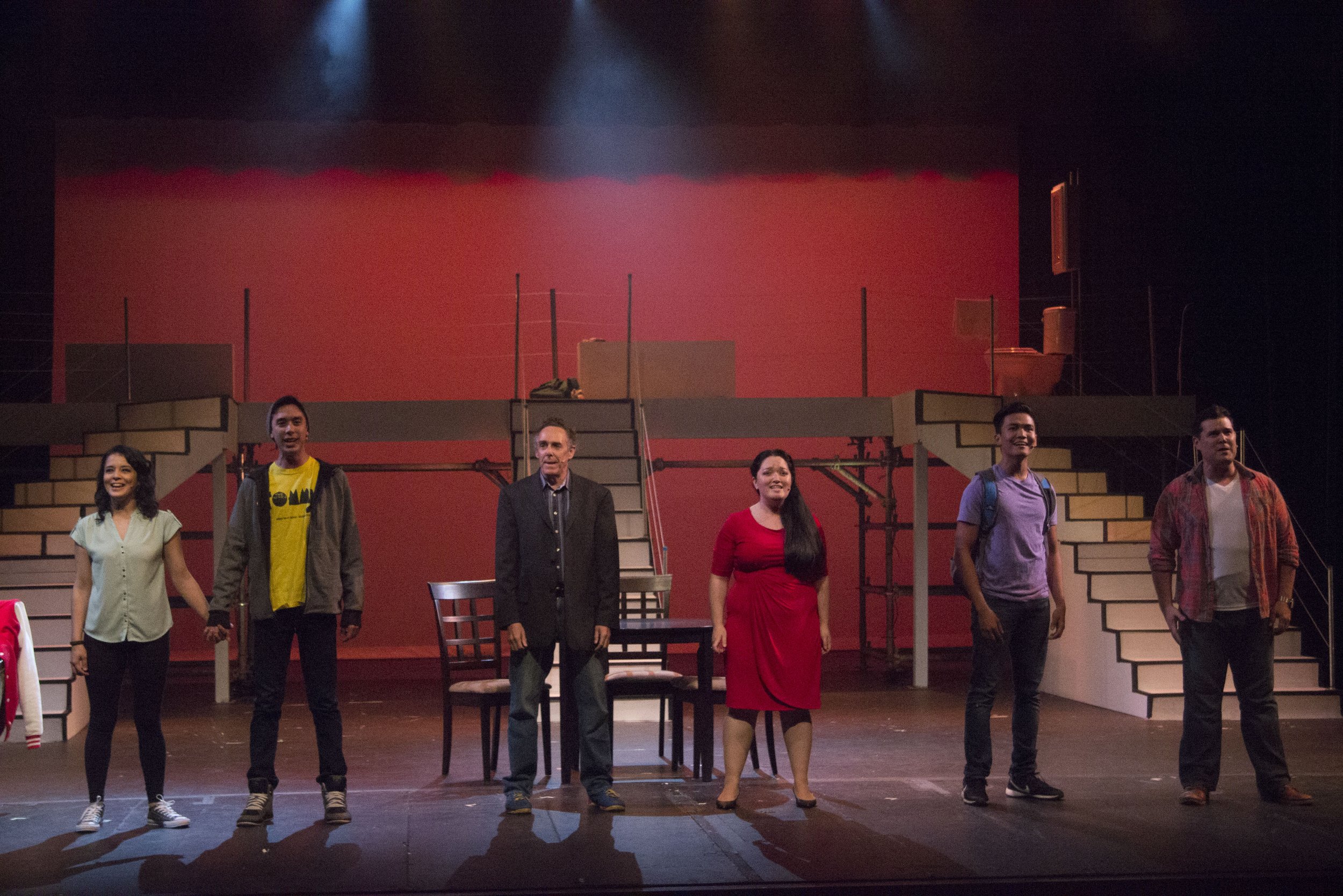 NEXT TO NORMAL a rock musical at Mamiya Theatre in Honolulu, Hawaii. Six people on stage in a theater, holding hands and smiling, with a set of stairs and a table in the background, part of a theatrical performance.