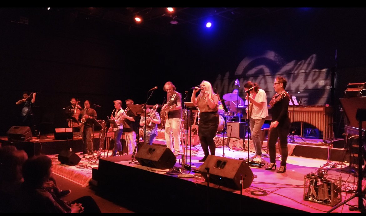 THE LIVING: A BENEFIT CONCERT BY BENNY RIETVELD OF SANTANA AND MILES DAVIS. Live show at Manoa Valley Theatre with Cassie Favreau. A band performing live on stage with multiple musicians, singers, and instruments, under colorful stage lighting.