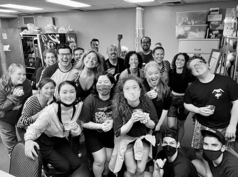 Backstage at Diamond Head Theatre in the Green Room after a rehearsal. Cheers! Group of people celebrating indoors, some wearing masks, smiling, holding drinks, in a room with shelves and a vending machine in the background.