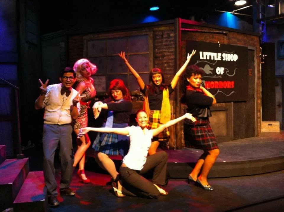 LITTLE SHOP OF HORRORS a comedic musical at Manoa Valley Theatre in Honolulu, Hawaii. Group of six people on a stage with a sign that reads 'Little Shop of Horrors' in the background. They are posing with cheerful and playful expressions.
