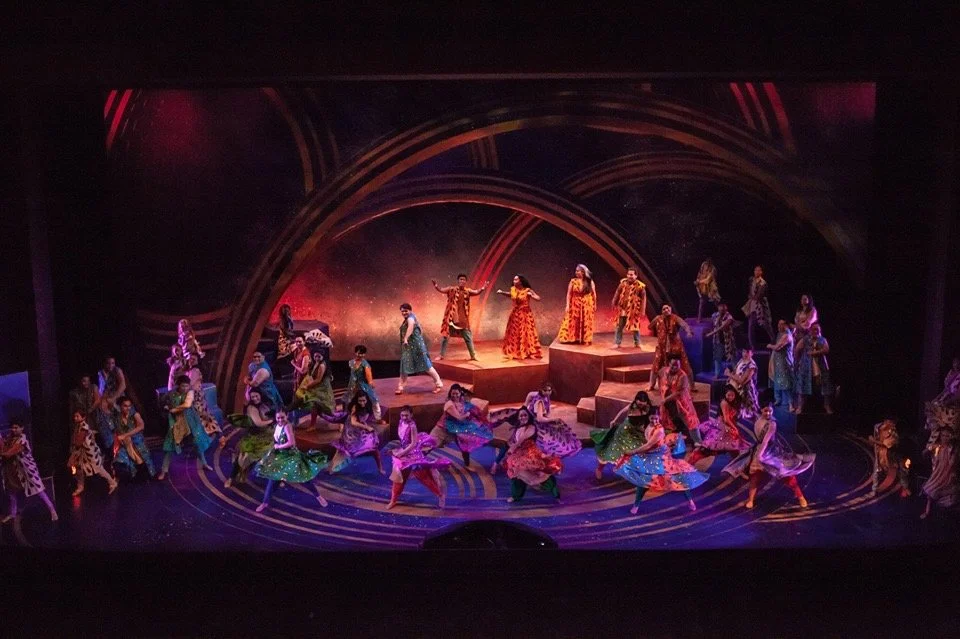 CHILDREN OF EDEN a Broadway musical by I'm A Bright Kid Foundation at Paliku Theatre. A stage performance with large group of dancers dressed in colorful costumes, performing in front of a vibrant, illuminated backdrop with artistic circular designs.