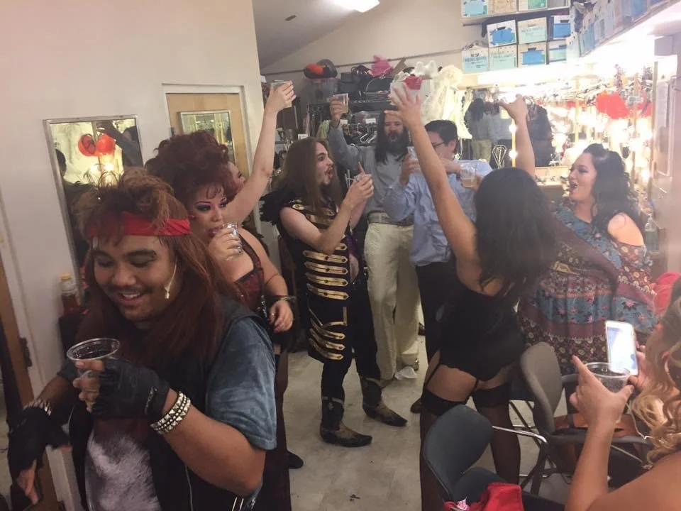 ROCK OF AGES a rock musical at Manoa Valley Theatre. Group of actors backstage celebrating after live show, dancing and holding drinks in a lively indoor setting.
