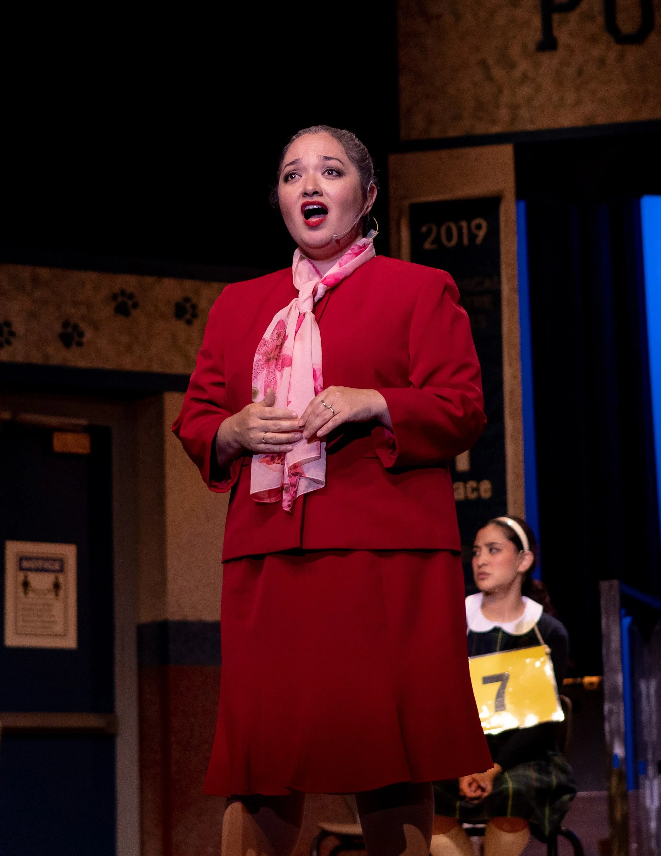 THE 25TH ANNUAL PUTNAM COUNTY SPELLING BEE a comedy musical at Manoa Valley Theatre in Honolulu, Hawaii. A woman in a red suit and pink scarf singing on stage while a young girl in a black dress with a yellow number tag listens in the background.