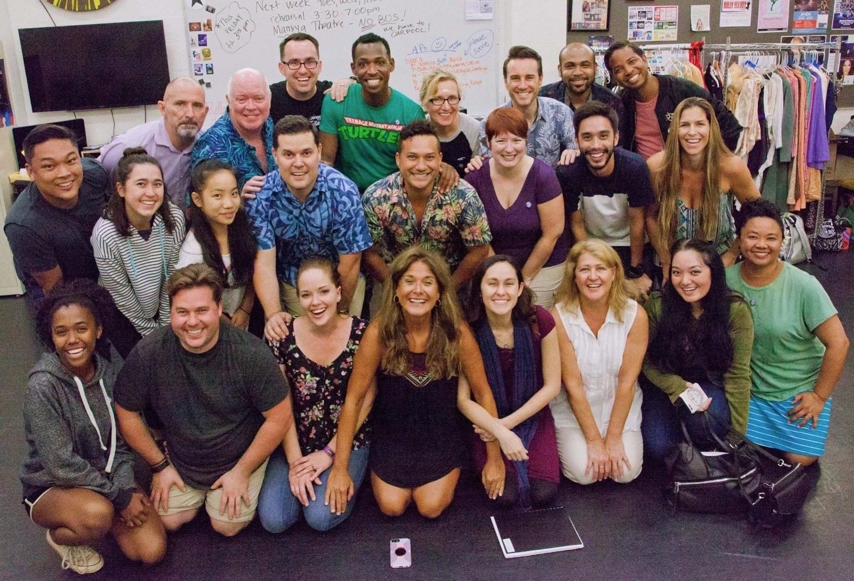 RISE UP: A BENEFIT CONCERT BY MELINA LILLIOS at Paliku Theatre in Kaneohe, Hawaii. A large group of people, smiling and posing together, in a classroom or community space with a whiteboard, television, and clothing racks in the background.