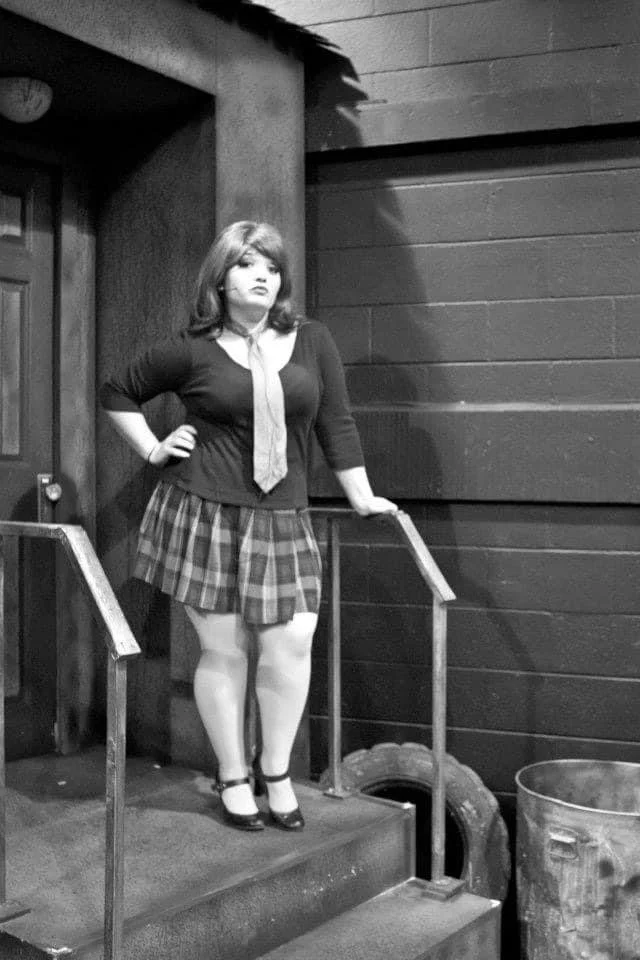 LITTLE SHOP OF HORRORS a comedic musical at Manoa Valley Theatre. A woman with shoulder-length hair, wearing a black top, plaid skirt, and high heels, standing on stairs next to a railing in an indoor setting.
