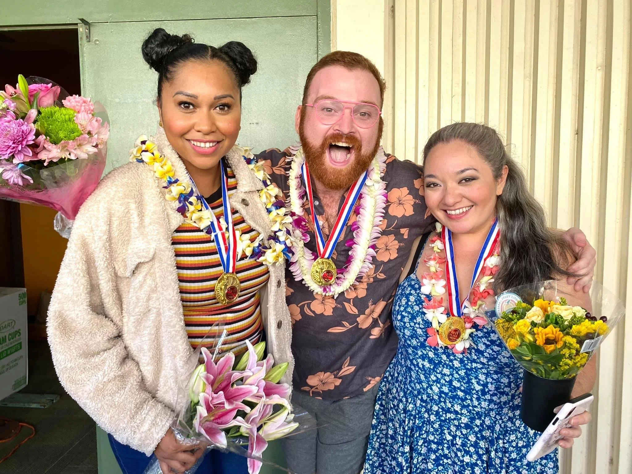 THE 25TH ANNUAL PUTNAM COUNTY SPELLING BEE a comedy musical at Manoa Valley Theatre in Honolulu, Hawaii. Three people with medals around their necks smiling, holding flowers, celebrating indoors against a yellow wooden wall.