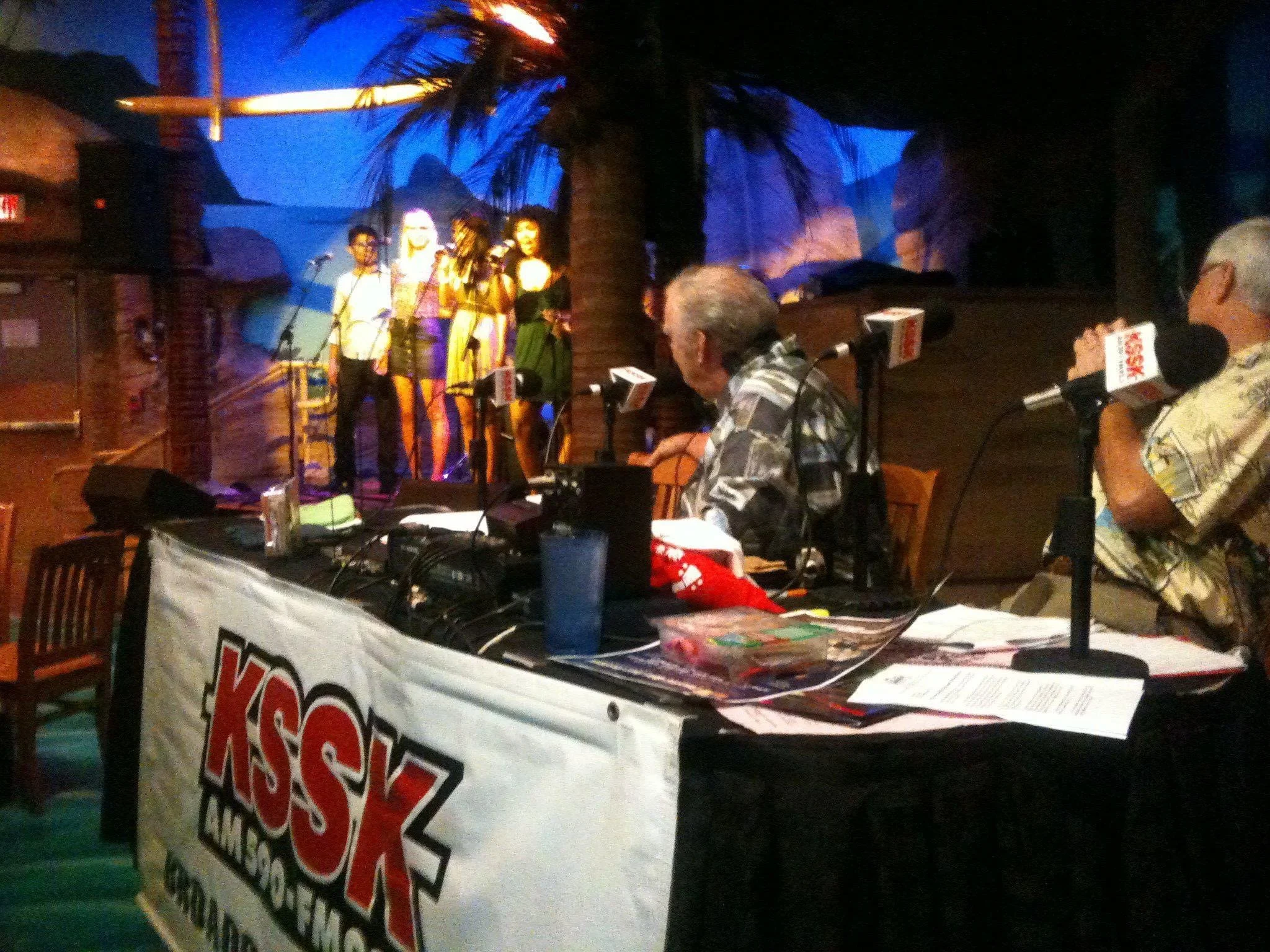 LITTLE SHOP OF HORRORS a comedic musical at Manoa Valley Theatre. Radio show. Four singers perform on a stage with a tropical decor, including palm trees, while four radio hosts sit at a table with KSSK banners and microphones in the foreground.