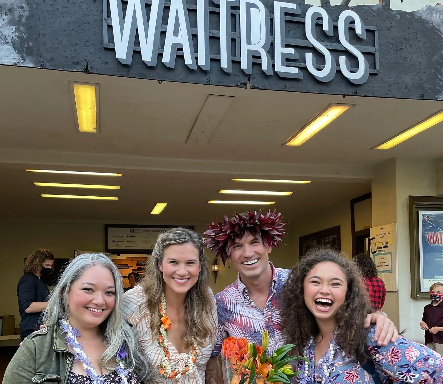 Four people smiling and posing together in front of a theatre entrance named "WATRESS," with some wearing leis and holding flowers, celebrating an event or performance.