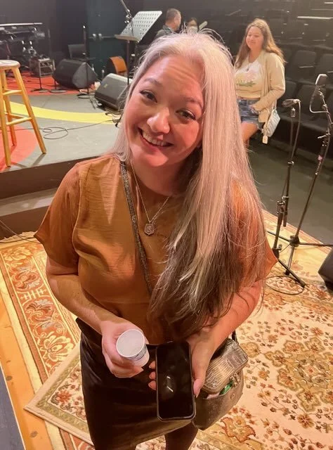 THE LIVING: A BENEFIT CONCERT BY BENNY RIETVELD OF SANTANA. A woman with long gray hair smiling at the camera, holding a water bottle, a phone, and a purse, on a stage with musical equipment and another woman sitting in the background.