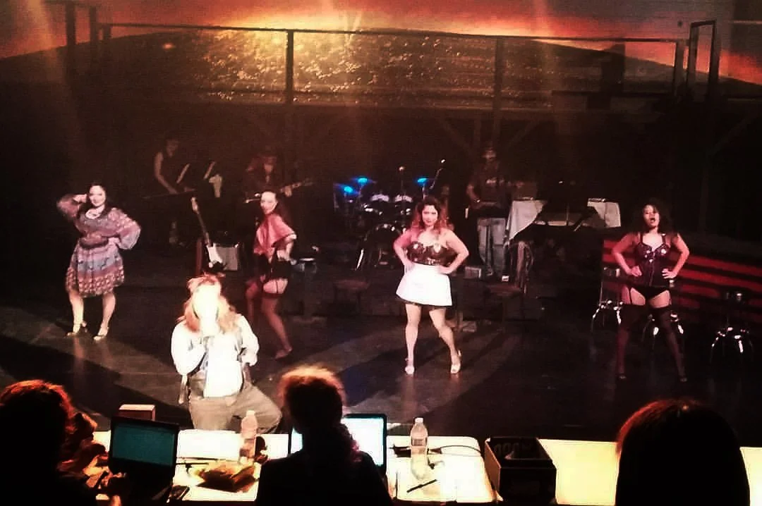 ROCK OF AGES a rock musical at Manoa Valley Theatre. A stage performance featuring six women in colorful costumes, with three women dancing in the front and three in the background, accompanied by a live band with a drummer and guitarists.