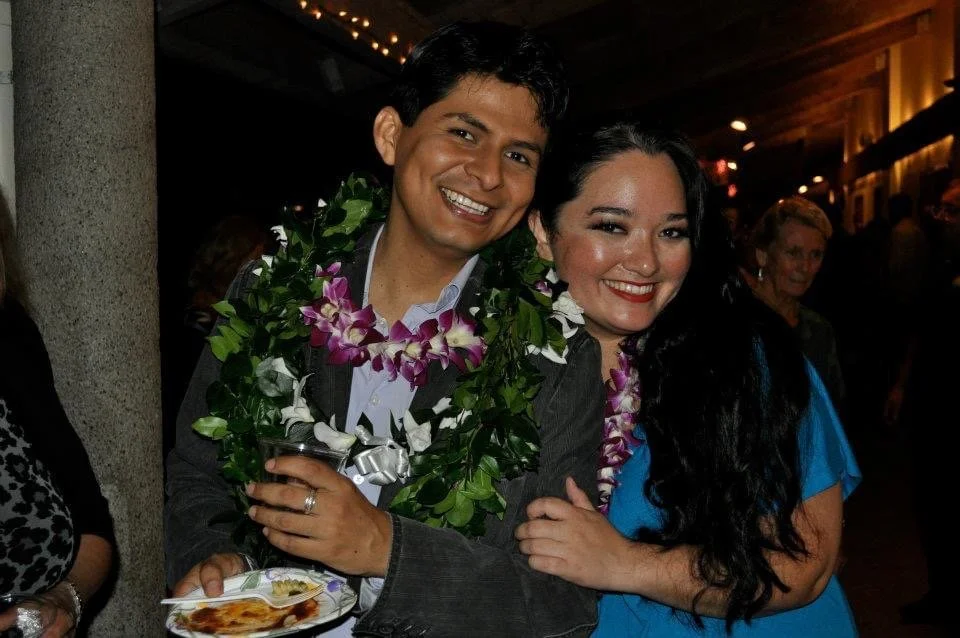 LITTLE SHOP OF HORRORS a comedic musical. Seymour and Crystal, smiling and embracing at a celebration event. The man is wearing a flower garland with purple and white flowers. The background suggests an indoor venue with warm lighting.