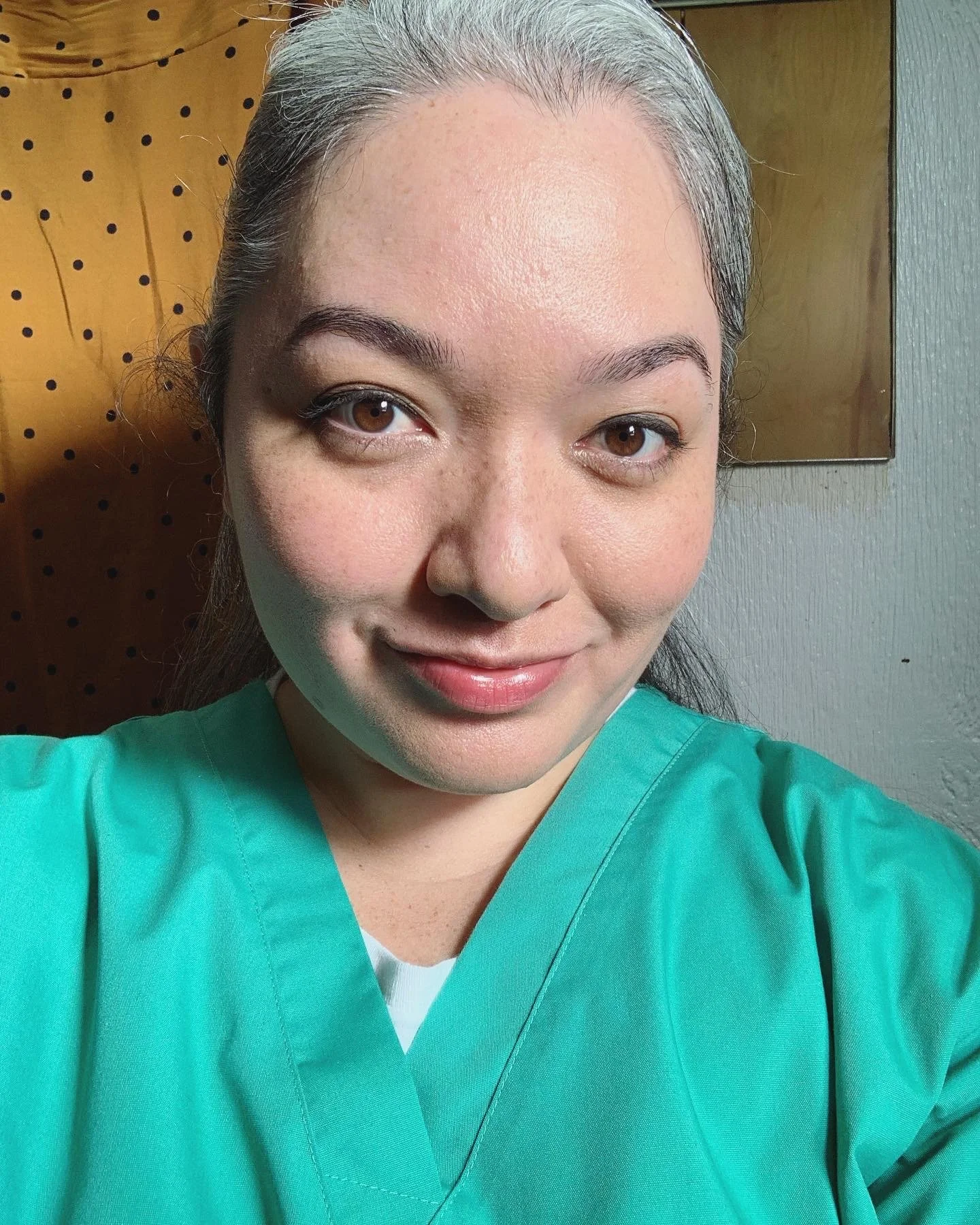 On set filming for MAGNUM P.I. with CBS on the studio lot. A SAG-AFTRA actor in green scrubs taking a selfie, smiling with brown eyes, standing indoors near a wooden wall and gray painted wall.