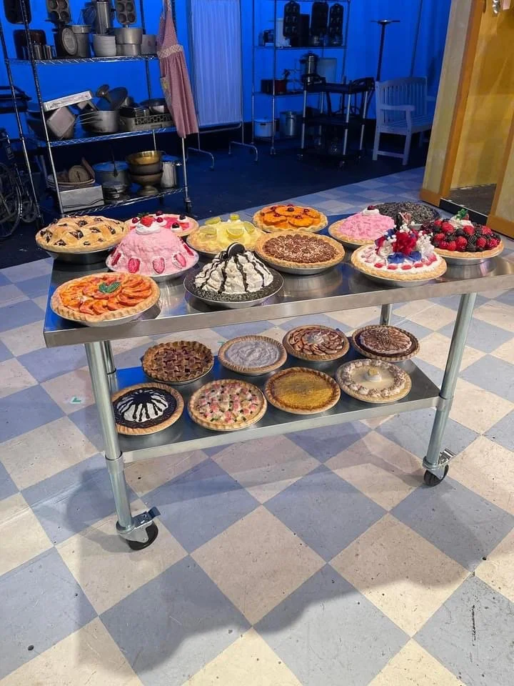 A stainless steel cart with two shelves displaying a variety of decorated pies and cakes in a commercial kitchen or bakery setting.