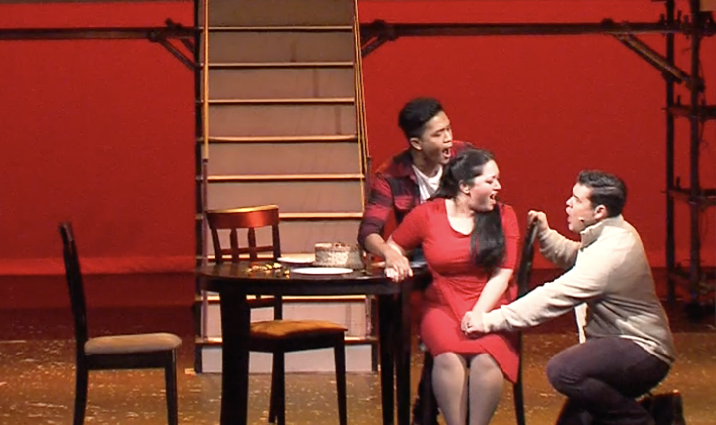 Three actors on stage in a theatrical play. Two are sitting, one kneeling, and one standing behind, all engaged in a lively scene. The stage has red walls, a staircase in the background, and a table with chairs, suggesting a domestic setting.