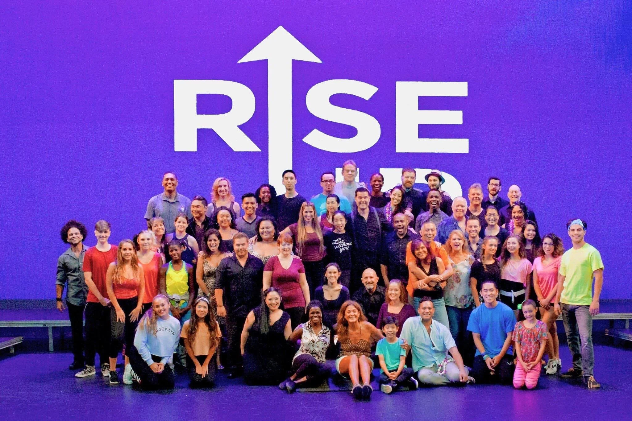 RISE UP: A BENEFIT CONCERT BY MELINA LILLIOS at Paliku Theatre in Kaneohe, Hawaii. Group photo of many people of various ages and ethnicities posing on stage with a large screen behind them displaying the words 'RISE UP' with an upward arrow.