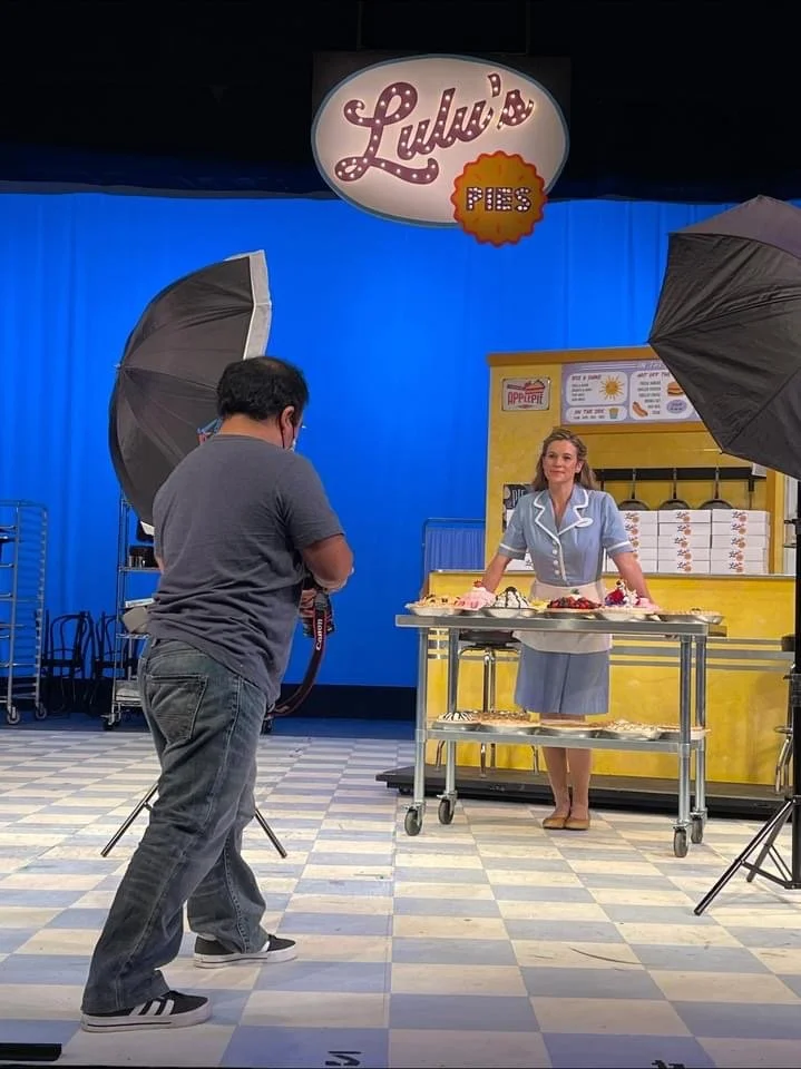 Photograph of a woman in a vintage-style blue dress standing behind a cake display on a cart, during a photoshoot or filming in a bakery-themed setting. There are tools and equipment like umbrellas for lighting, a man with a camera, and a large sign 