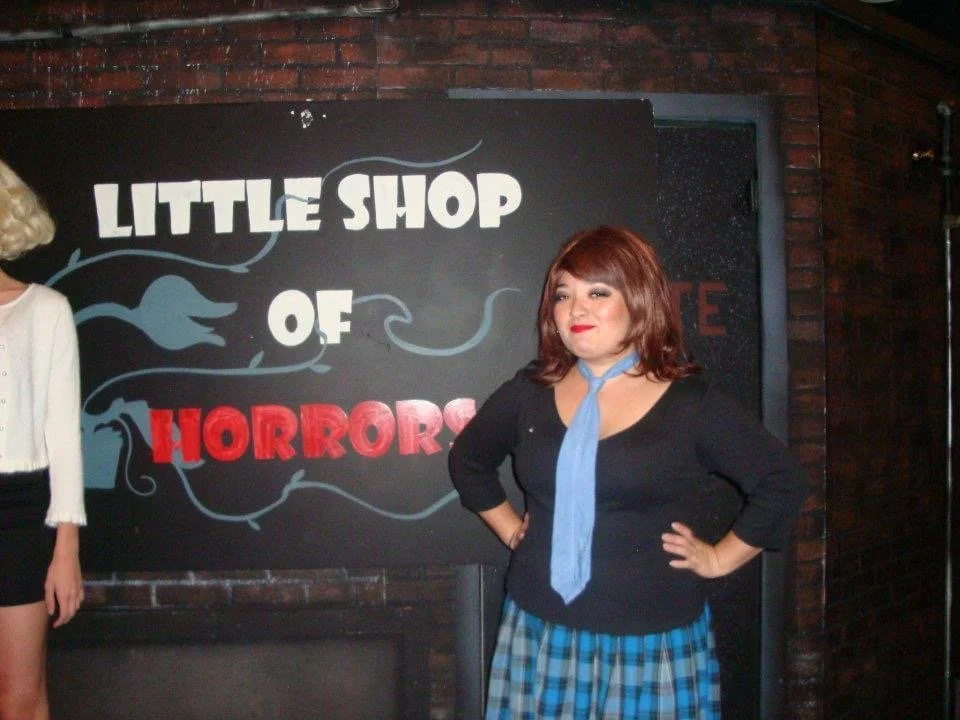 LITTLE SHOP OF HORRORS a comedic musical at Manoa Valley Theatre. Woman standing with hands on hips in front of a sign that reads 'Little Shop of Horrors' against a brick wall, with part of another person's arm and clothing visible on the left.