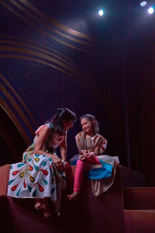CHILDREN OF EDEN a Broadway musical by I'm A Bright. Two young girls sitting on a stage, smiling and engaging in a conversation; one girl wearing a colorful dress with patterns, the other in a dress with bright pink leggings. Stage lighting overhead.
