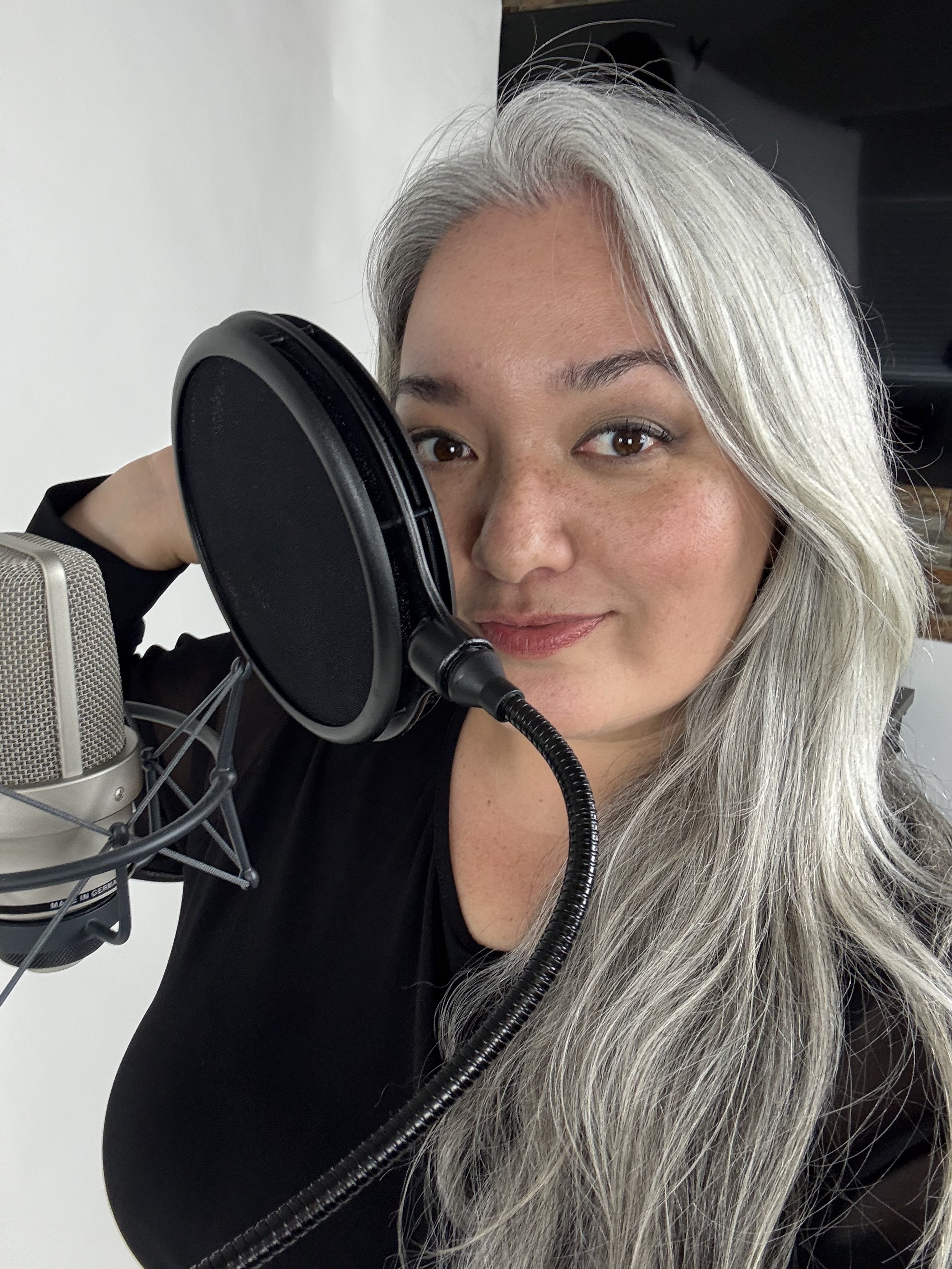Hawaii voiceover actress. A woman with long gray hair is speaking into a professional microphone with a pop filter, wearing a black shirt, in a recording studio.