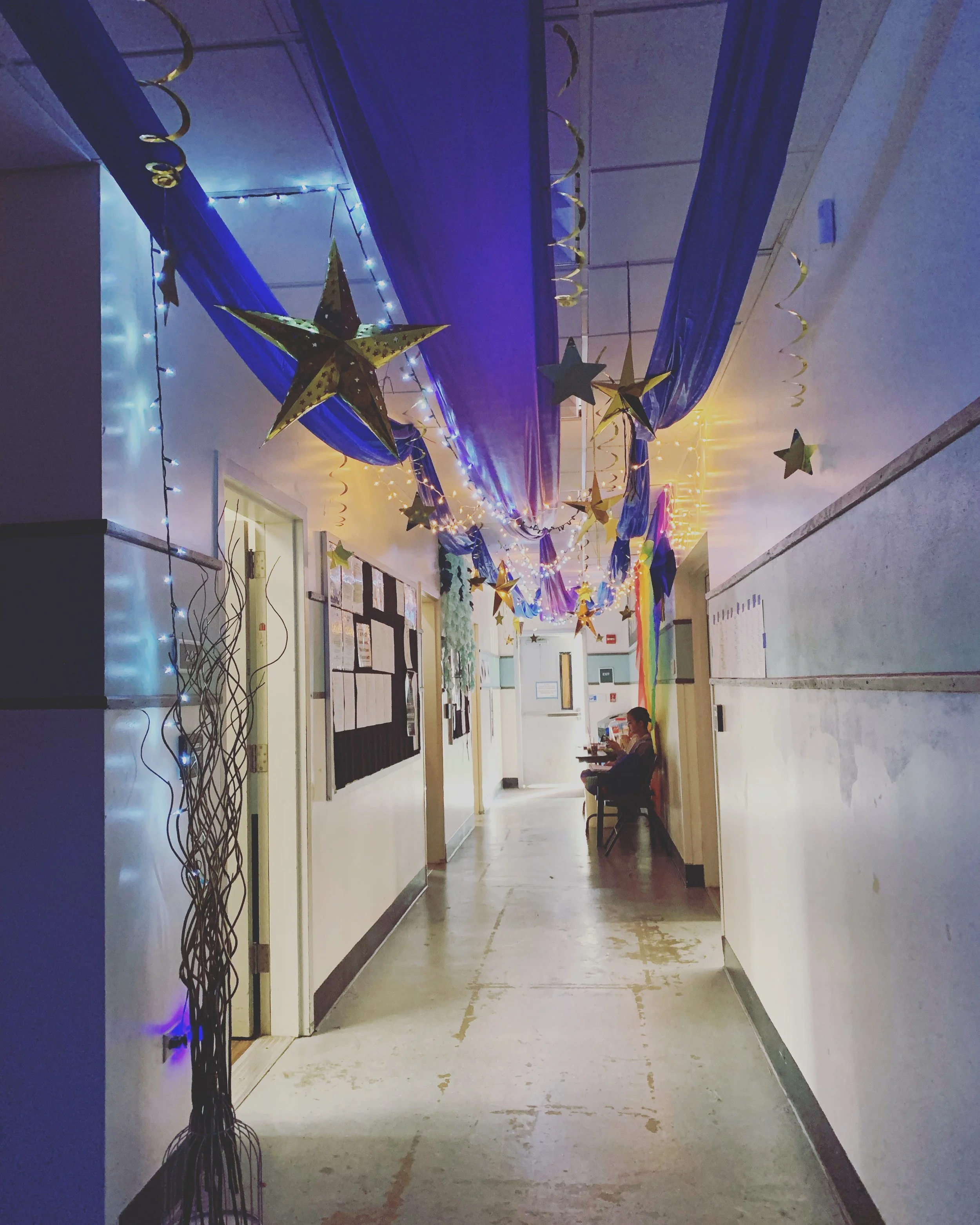 CHILDREN OF EDEN a Broadway musical by I'm A Bright Kid. Decorated school hallway with blue fabric draped along the ceiling, gold stars and string lights hanging, and a rainbow flag on the wall. Two people are sitting on chairs in the background.