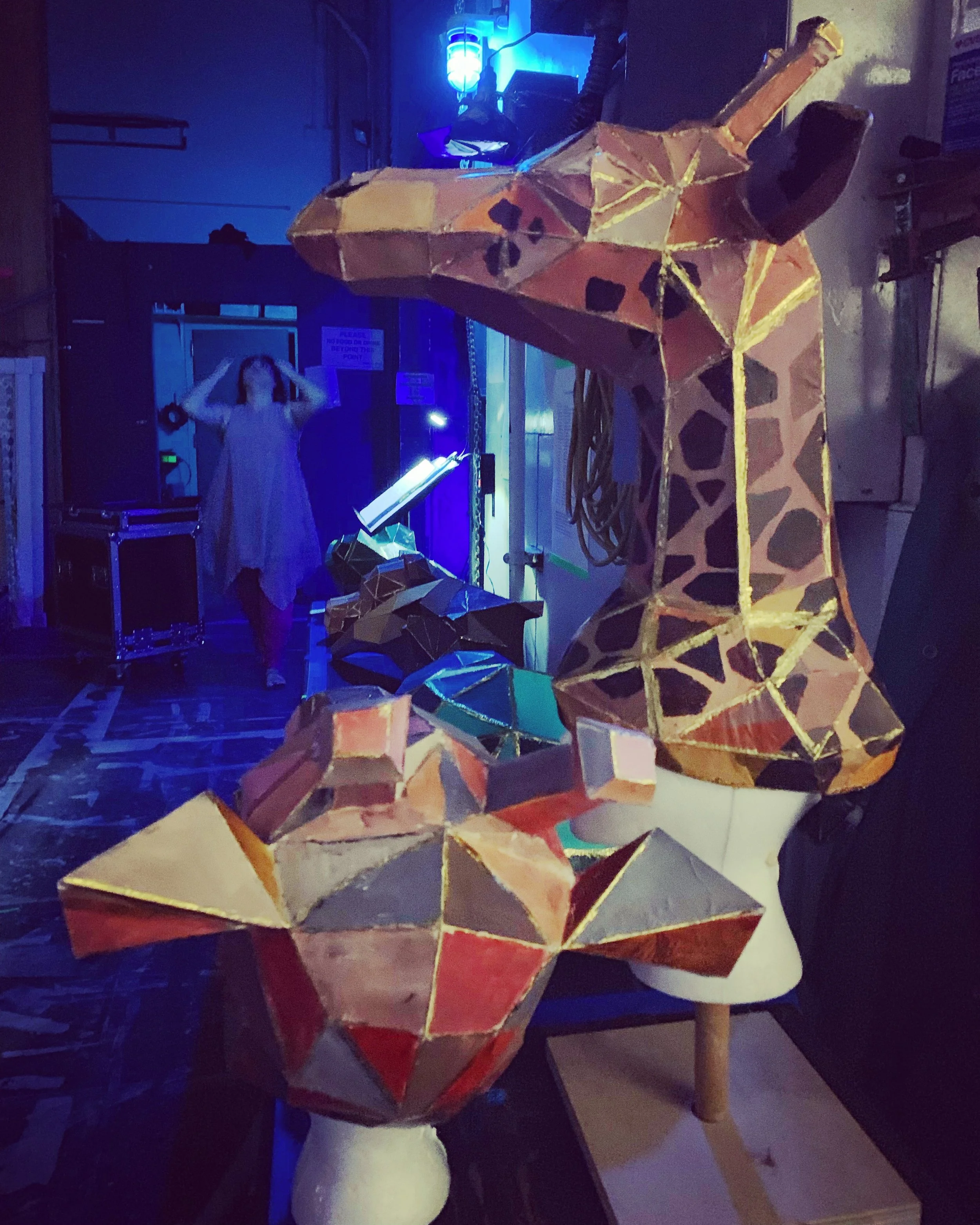 CHILDREN OF EDEN a Broadway musical by I'm A Bright Kid Foundation at Paliku Theatre. A large, geometric, paper-mâché giraffe sculpture on a mannequin head in a workshop, with a woman in the background adjusting her hair.