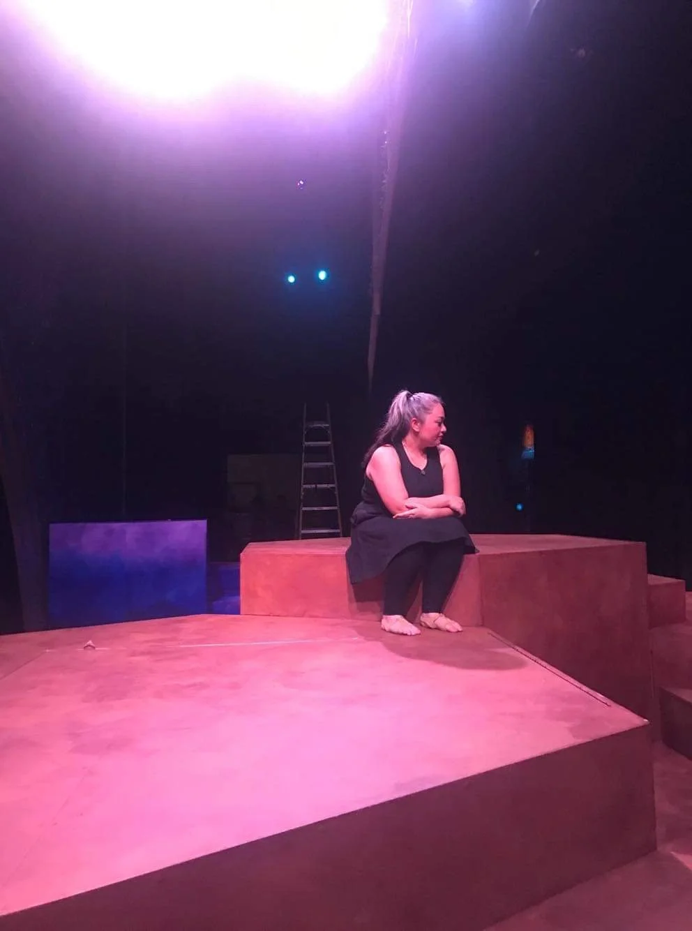 CHILDREN OF EDEN a Broadway musical by I'm A Bright Kid Foundation at Paliku Theatre. A woman sitting on a wooden stage with her arms crossed, looking to her left, with a ladder in the background and stage lights overhead.