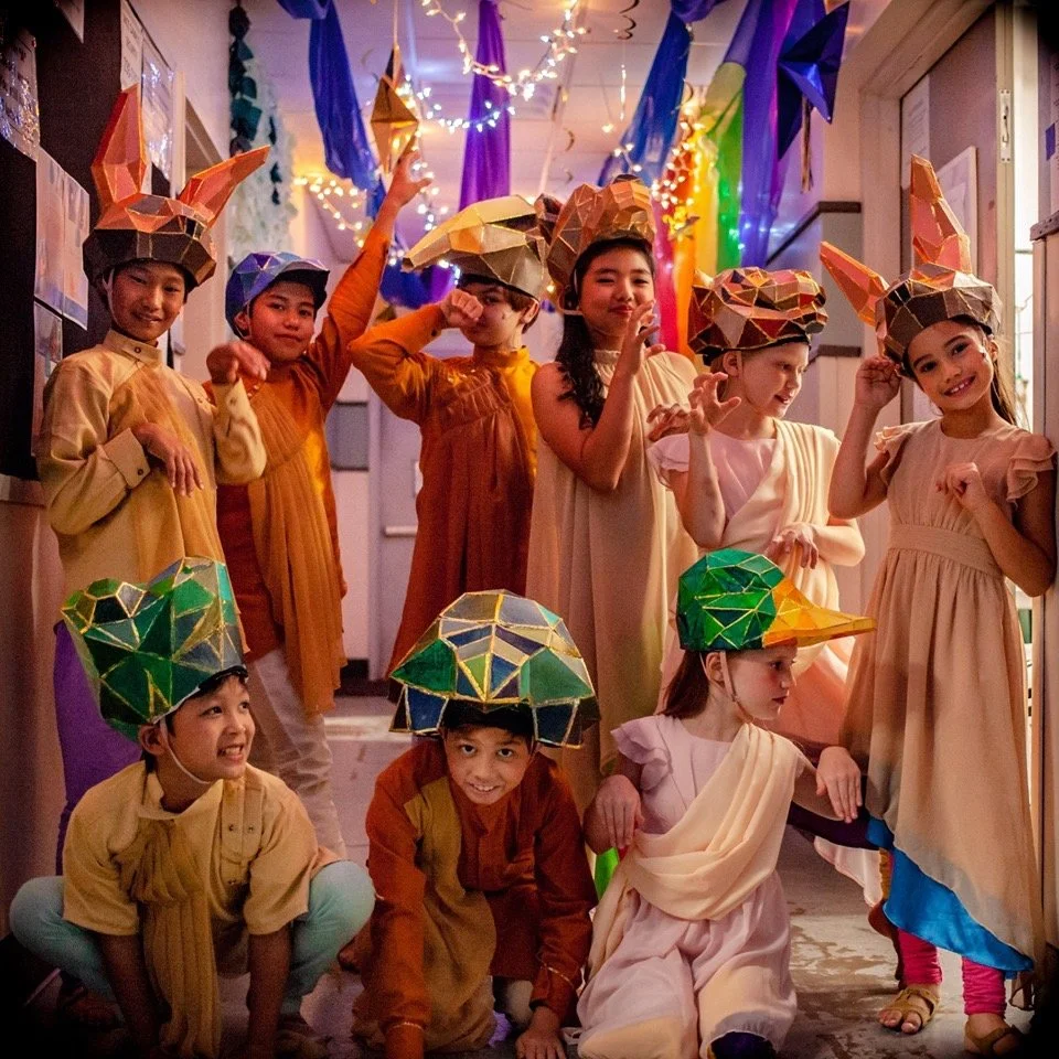 CHILDREN OF EDEN a Broadway musical by I'm A Bright Kid Foundation at Paliku Theatre. Group of children in colorful costumes and hats, celebrating indoors with festive decorations and string lights.