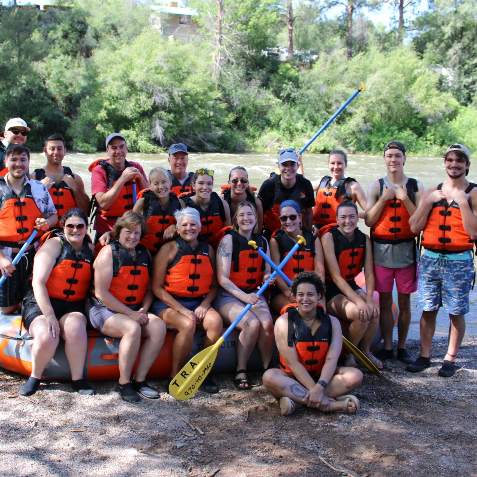 A group of people in orange life jackets standing on a riverbank with a raft, surrounded by trees, smiling for the camera after a rafting trip.