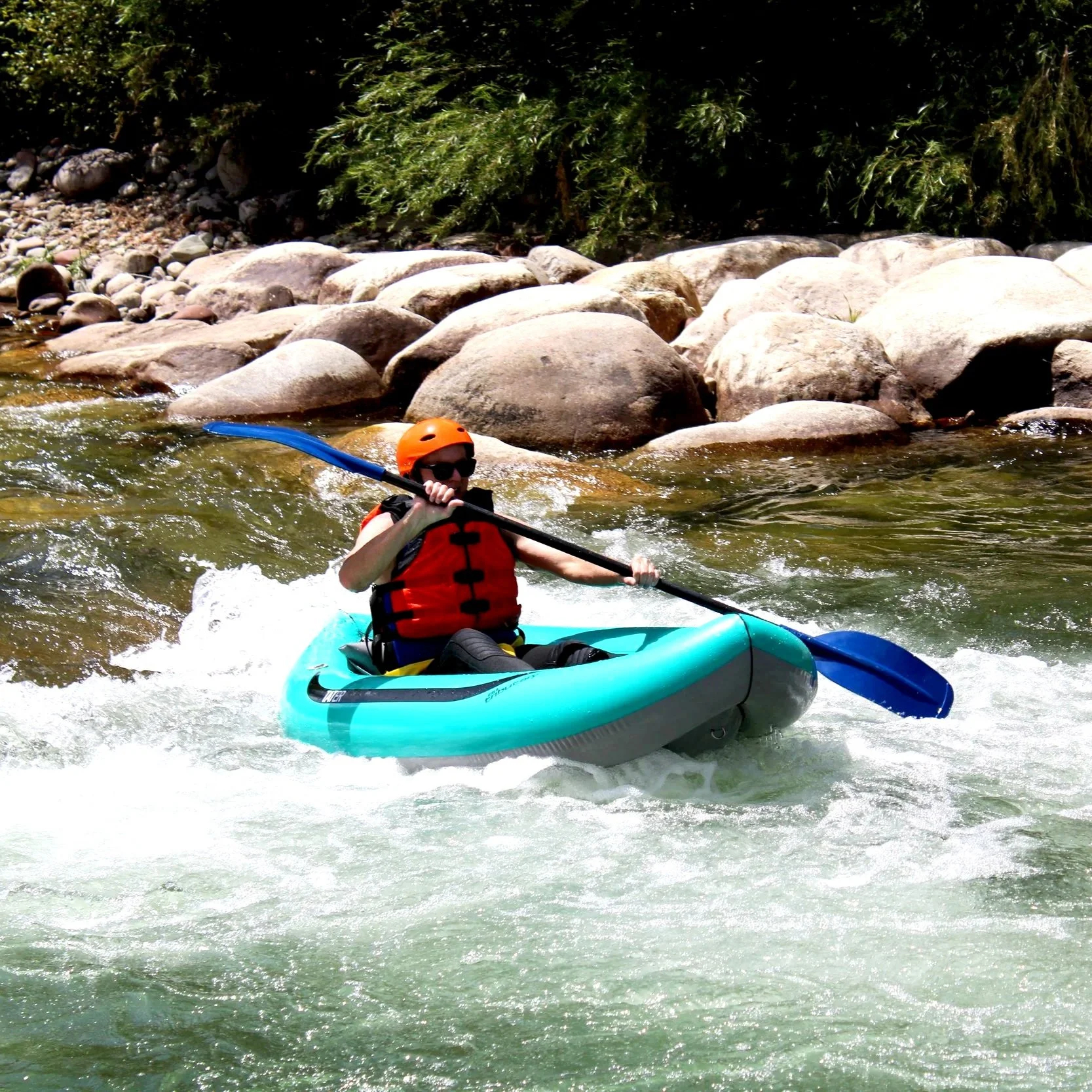 A person wearing an orange helmet, red life jacket, and sunglasses kayaking on a river surrounded by rocks and greenery.