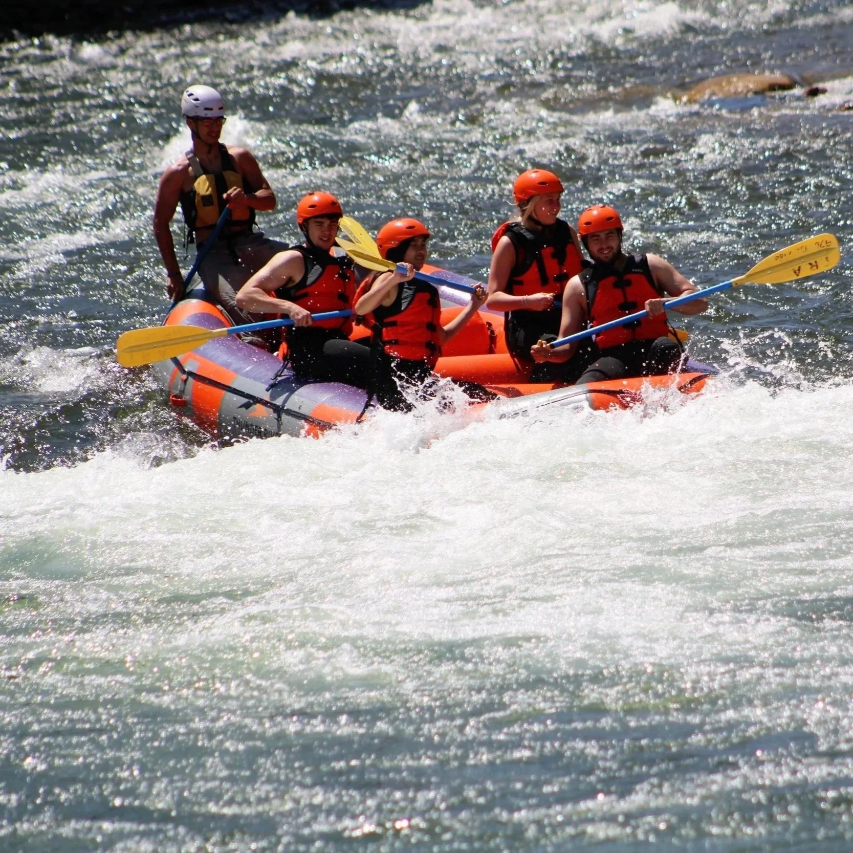 Five people white-water rafting on a river, wearing life jackets and helmets, with some holding paddles.