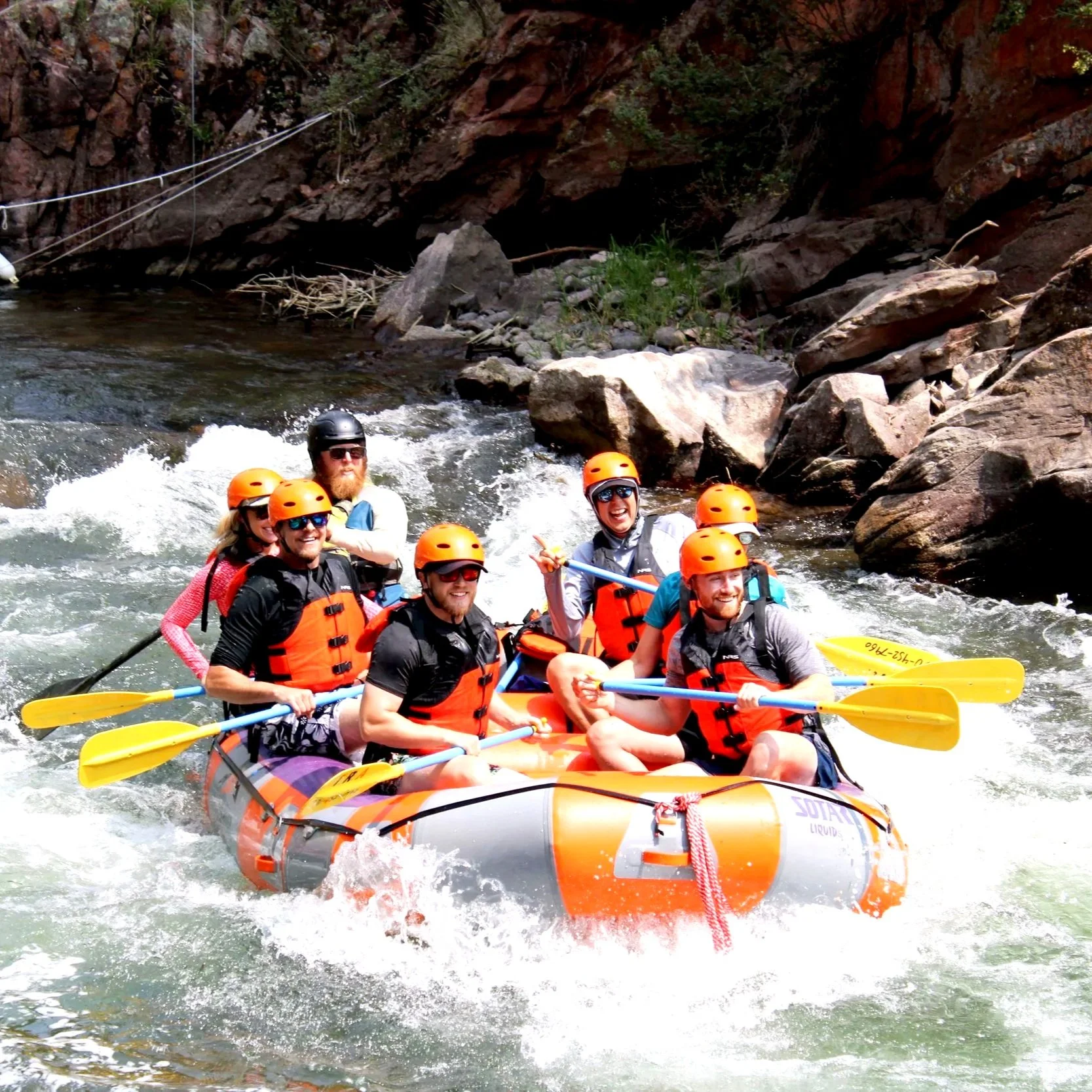 Group of seven people rafting on white water river, all wearing orange life jackets and helmets, smiling, and holding paddles.