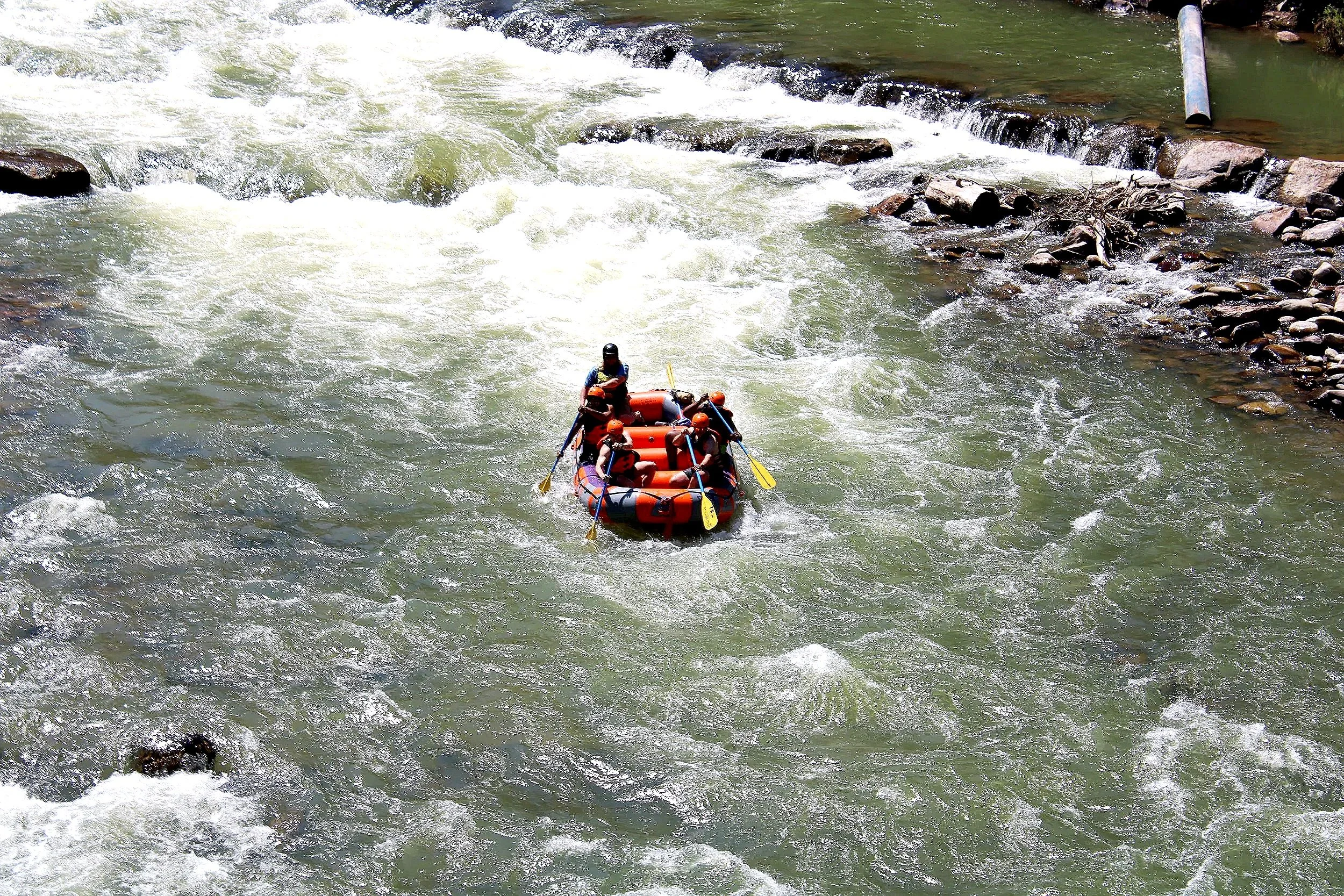 A group of six people in an orange raft navigating through white water rapids on a river, with rocky banks and some debris in the water.