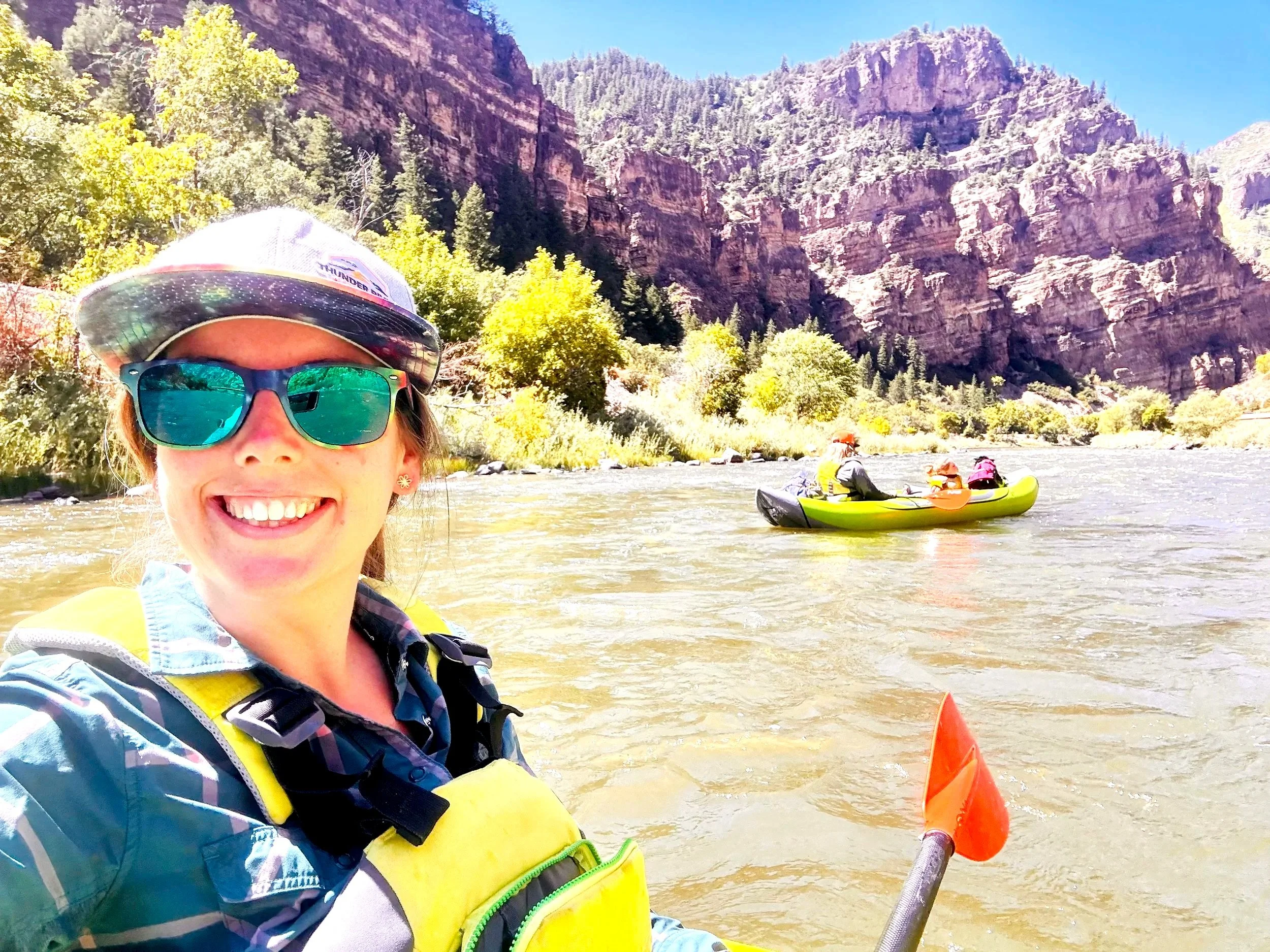 A woman smiling for a selfie while kayaking on a river, with a rocky canyon and trees in the background, and two people paddling a green kayak in the water.