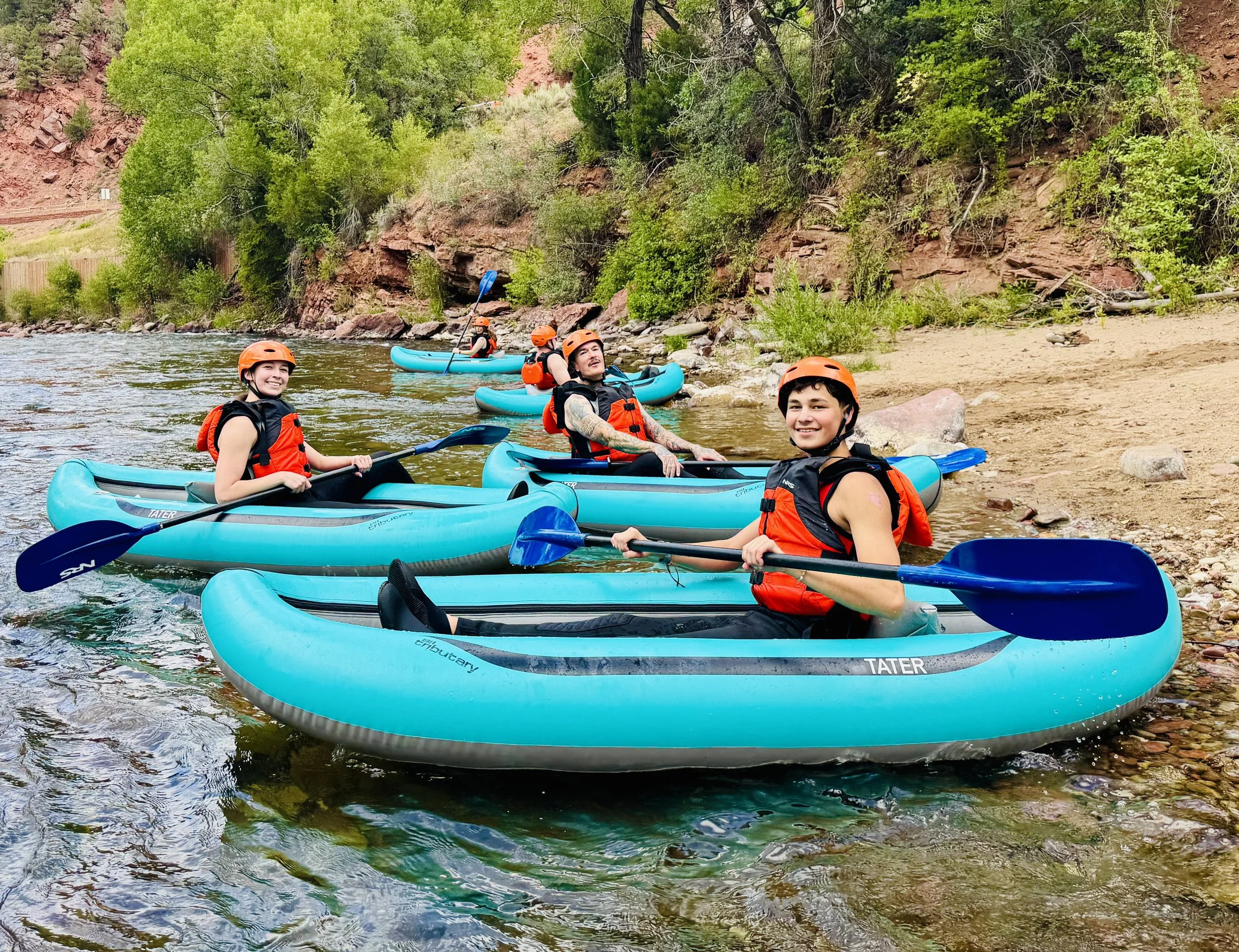 Group of five people in blue kayaks with orange life jackets and helmets, paddling on a river surrounded by trees and rocky cliffs.