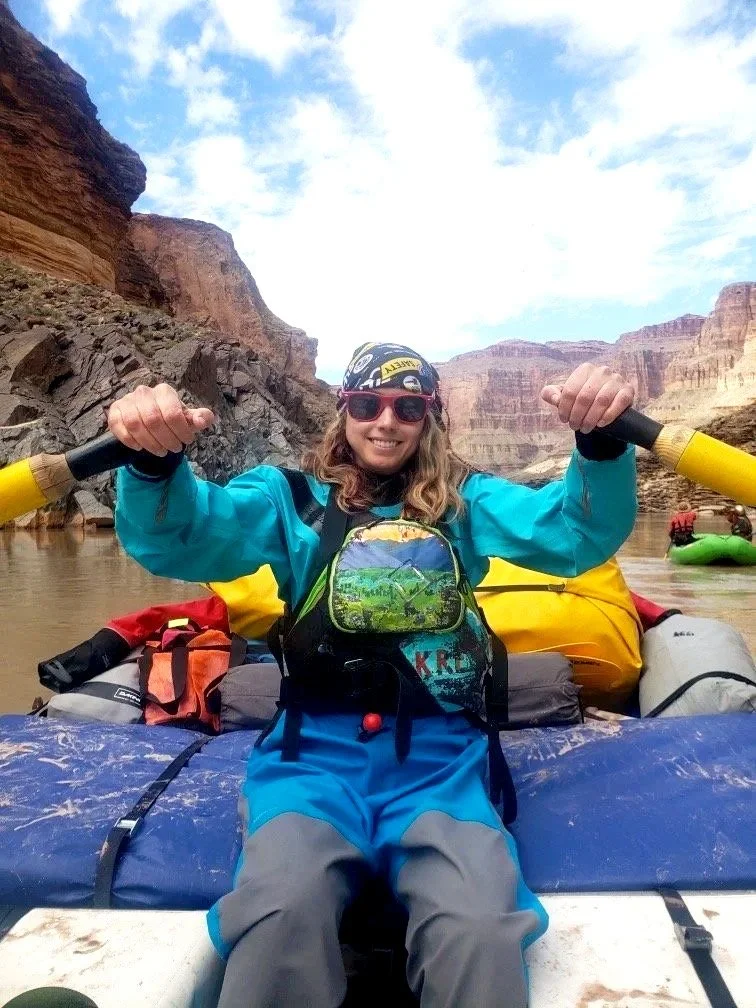 Woman in colorful outdoor gear smiling on a raft in a canyon with rocky cliffs and water.