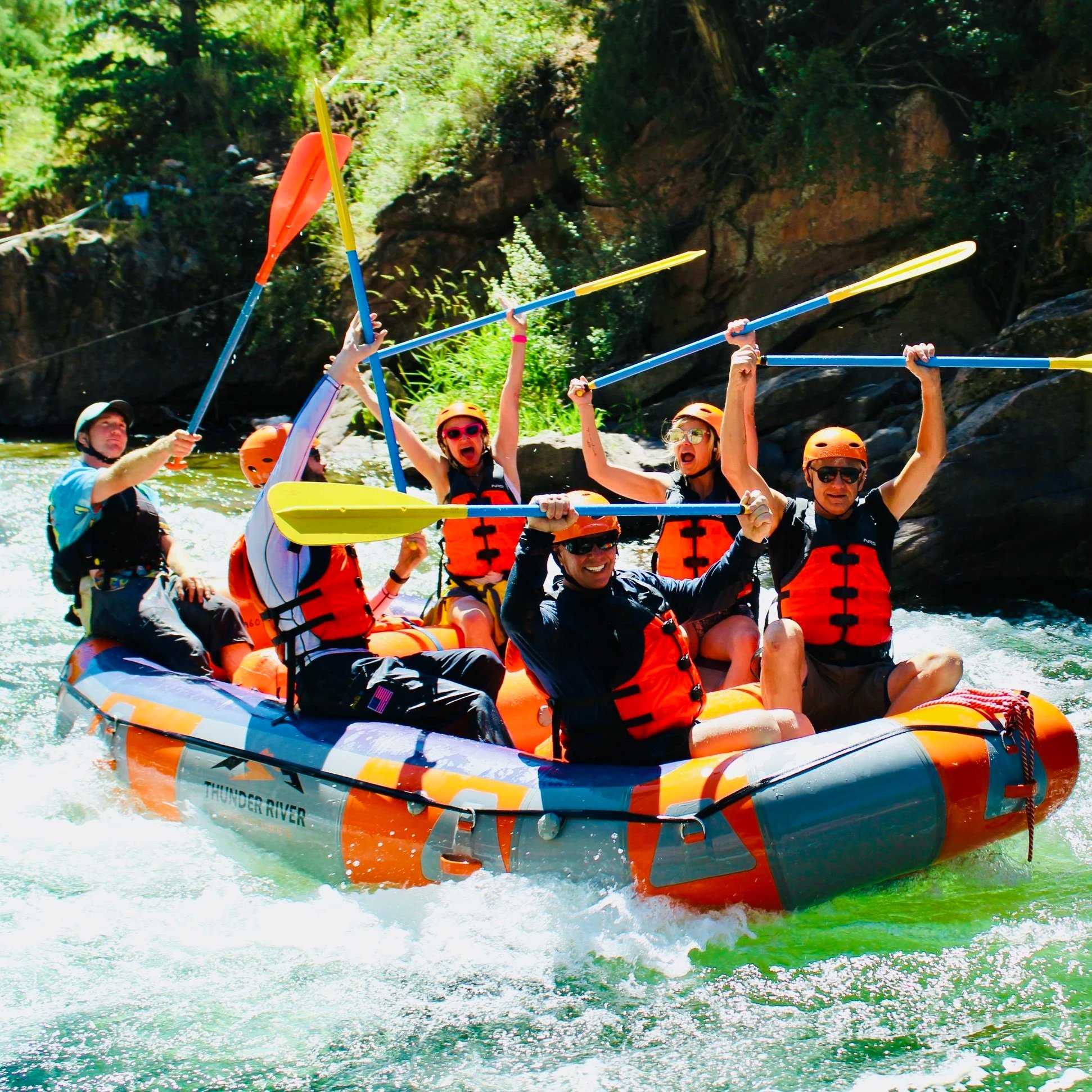 Group of six people white-water rafting on a river, wearing life jackets and helmets, holding paddles raised in cheers.