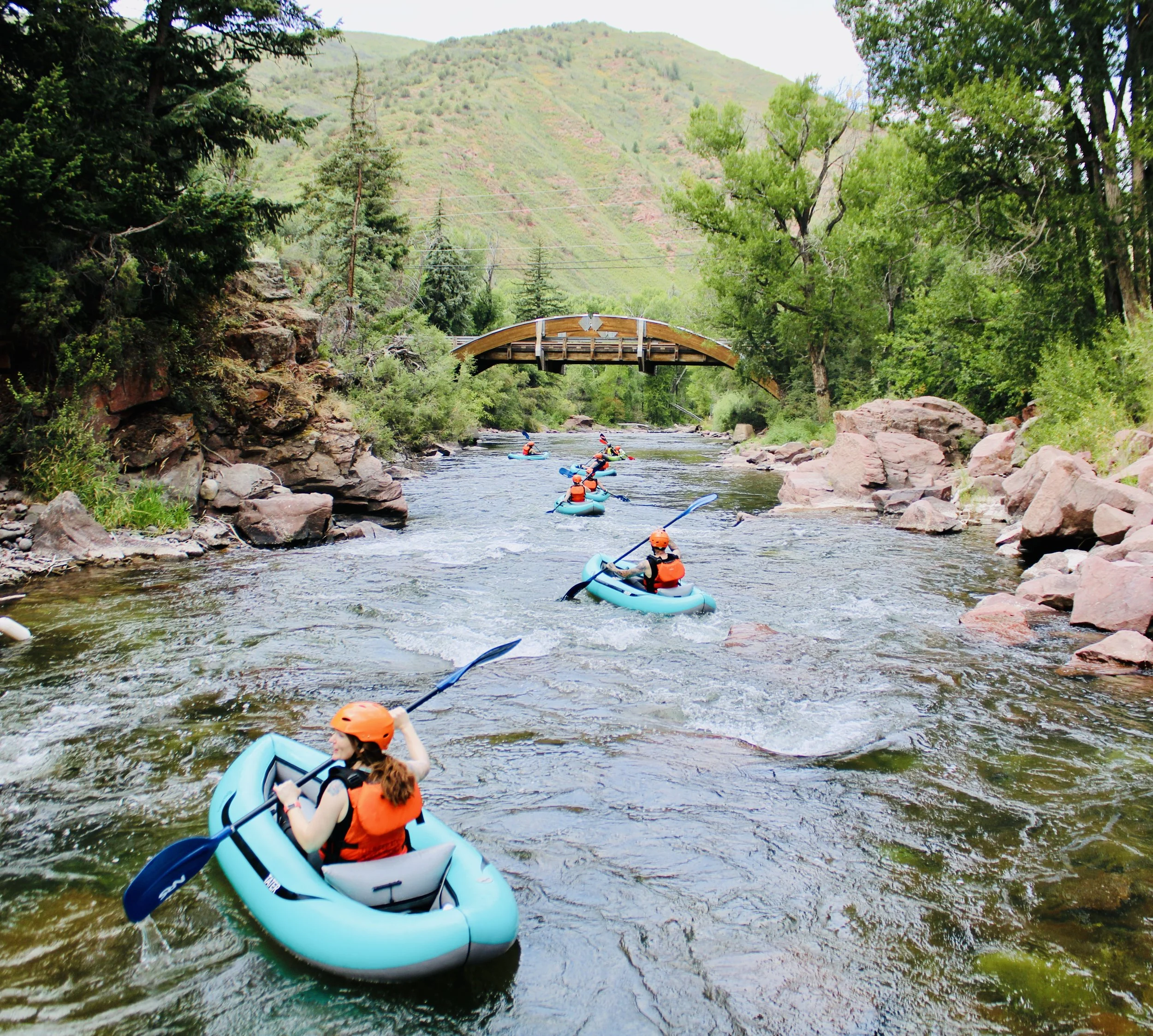 People kayaking on a river surrounded by rocks and green trees, with a bridge and hills in the background.