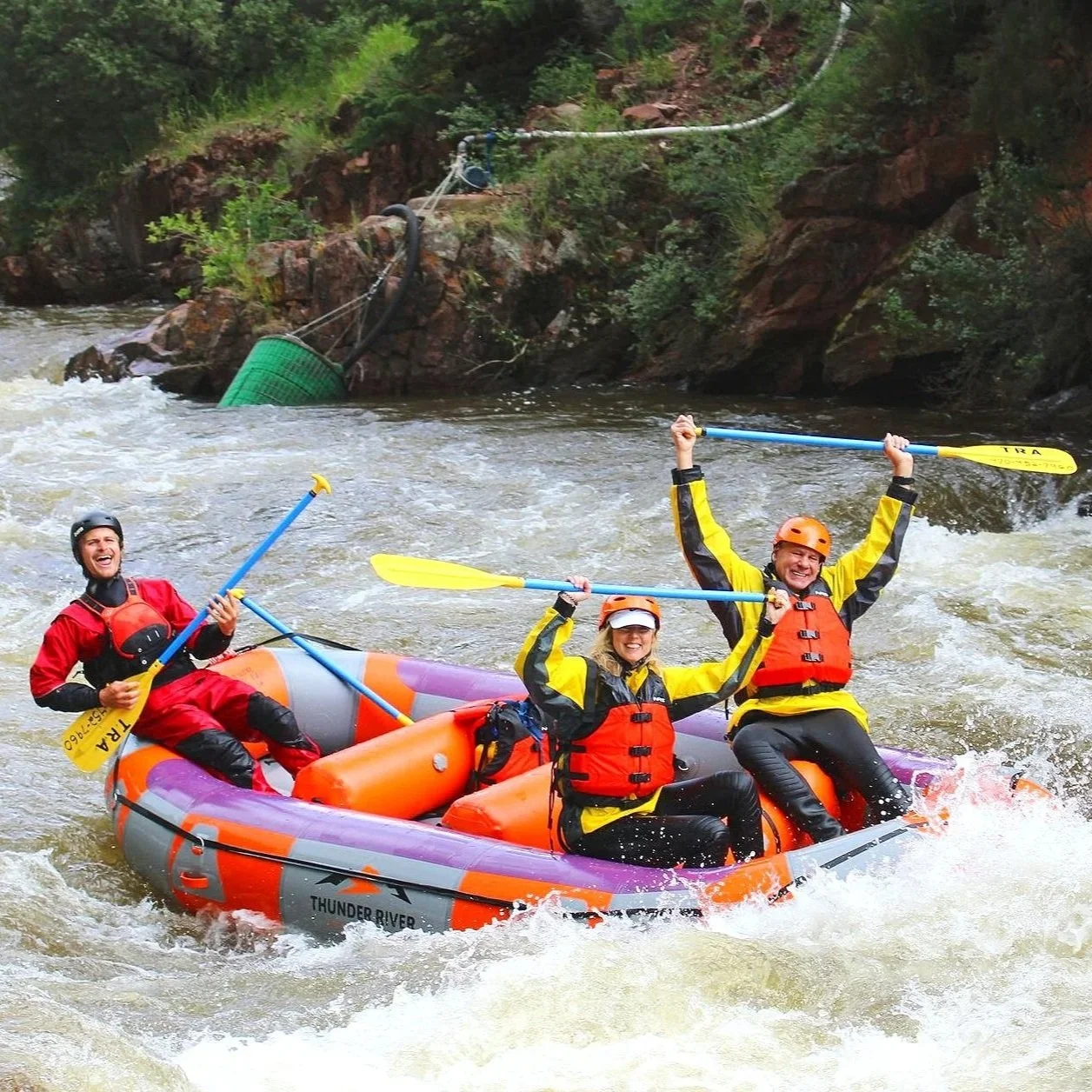Three people white-water rafting in an inflatable raft on a river, wearing life jackets and helmets, with two women and one man smiling and raising paddles in celebration.