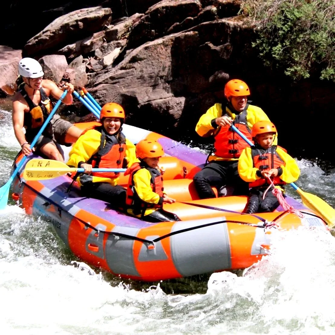Group of people white water rafting on a river, wearing helmets and life jackets, surrounded by rocks and water.
