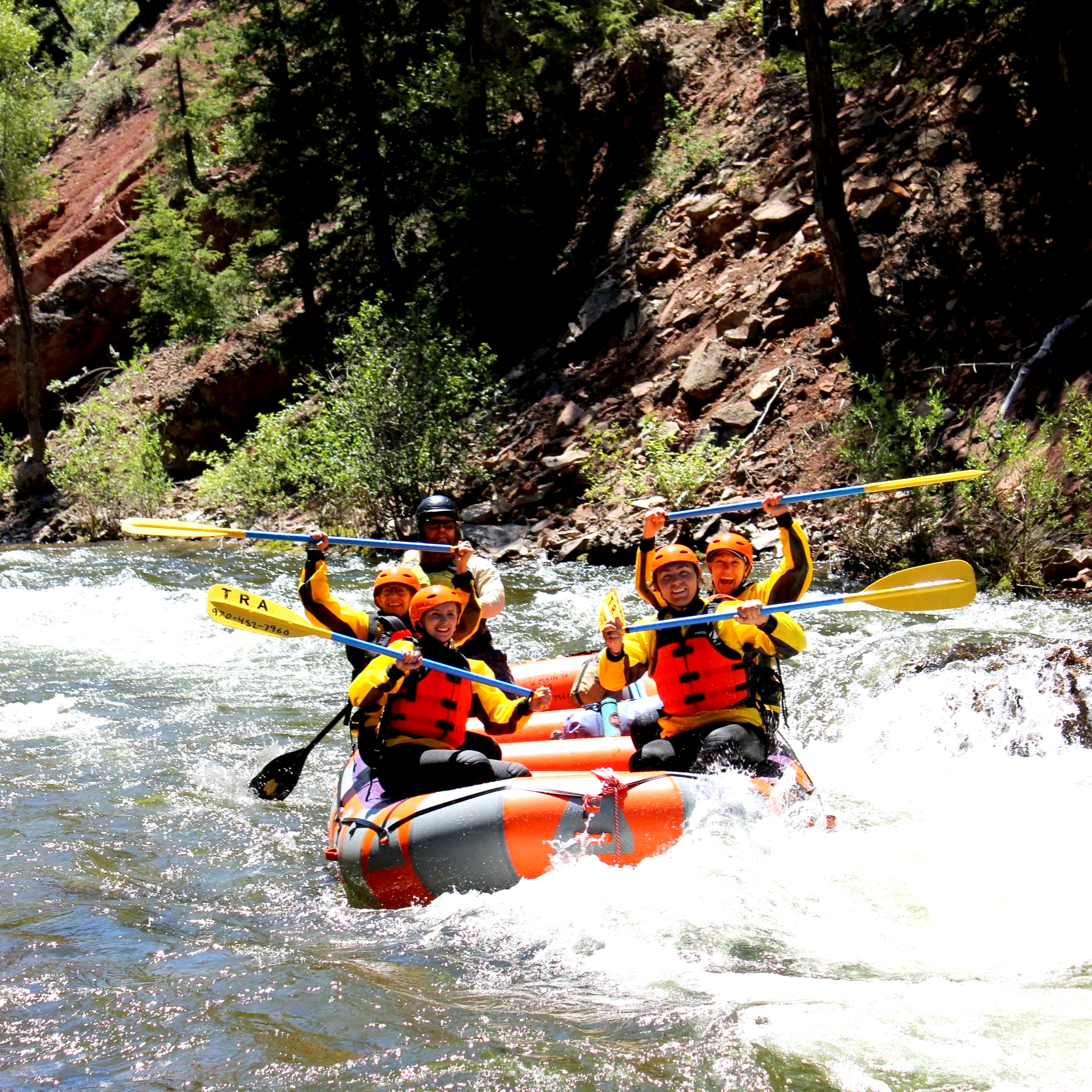 Group of people whitewater rafting on a river, wearing helmets and life jackets, with oars raised, surrounded by trees and rocks.