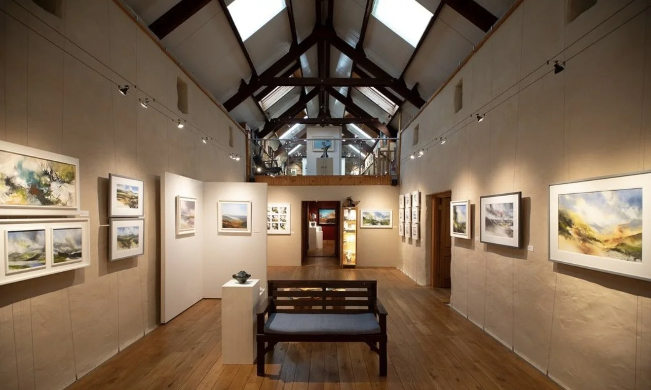 An art gallery with paintings hanging on beige walls, a wooden bench, and a skylight ceiling in a high-ceilinged space.