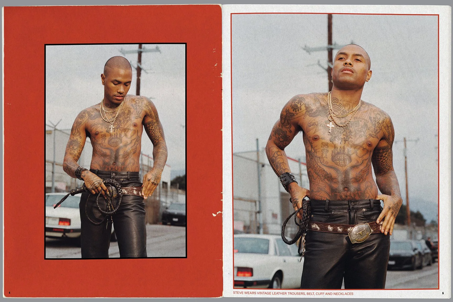 A shirtless man with multiple tattoos, wearing black leather trousers, a large belt with a decorative buckle, and multiple gold necklaces, stands outdoors in front of a chain-link fence and parked cars, with a slightly overcast sky.